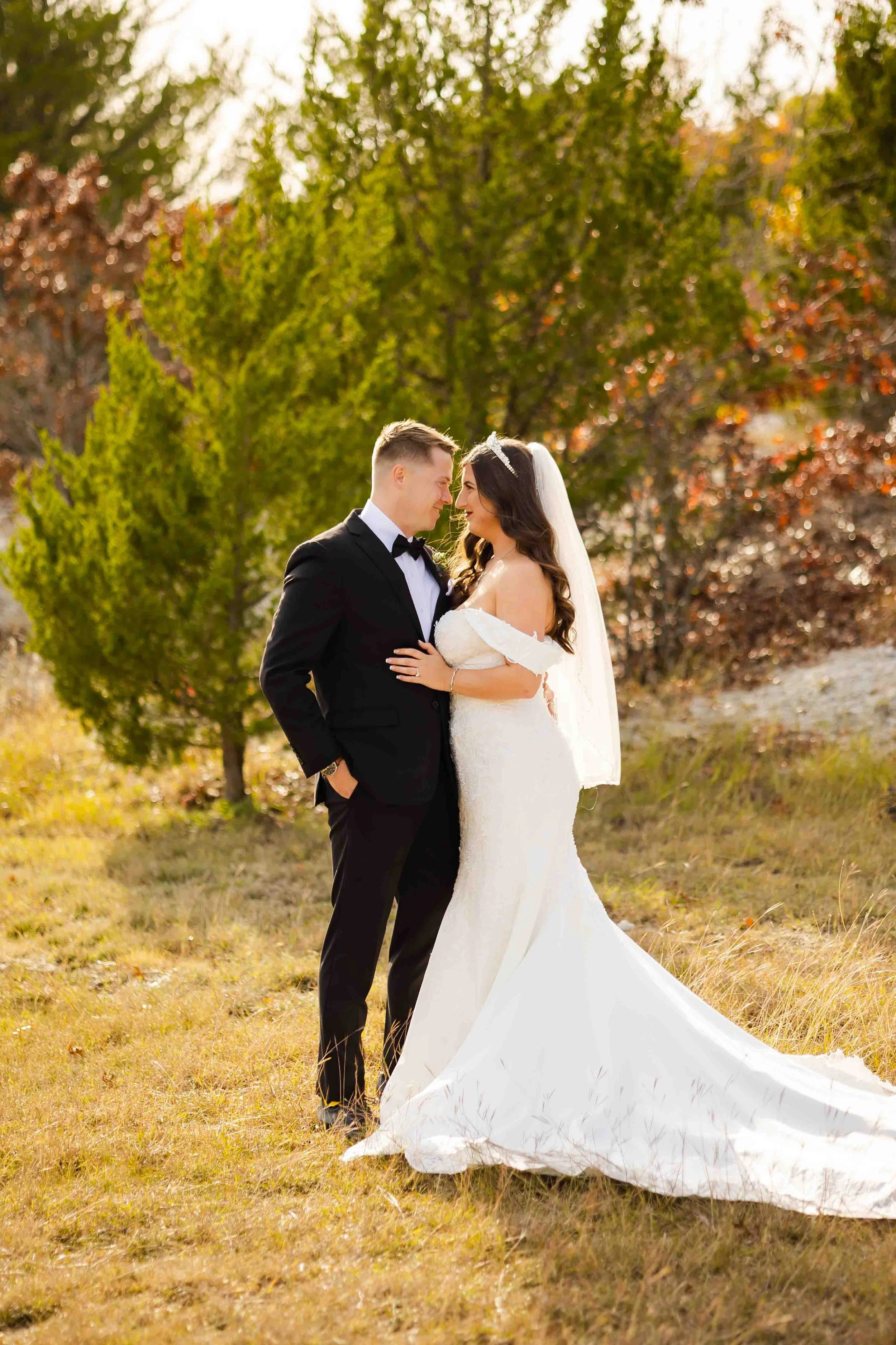 A bride and groom standing close together outdoors in a grassy area with trees and fall foliage in the background, during a wedding photoshoot.