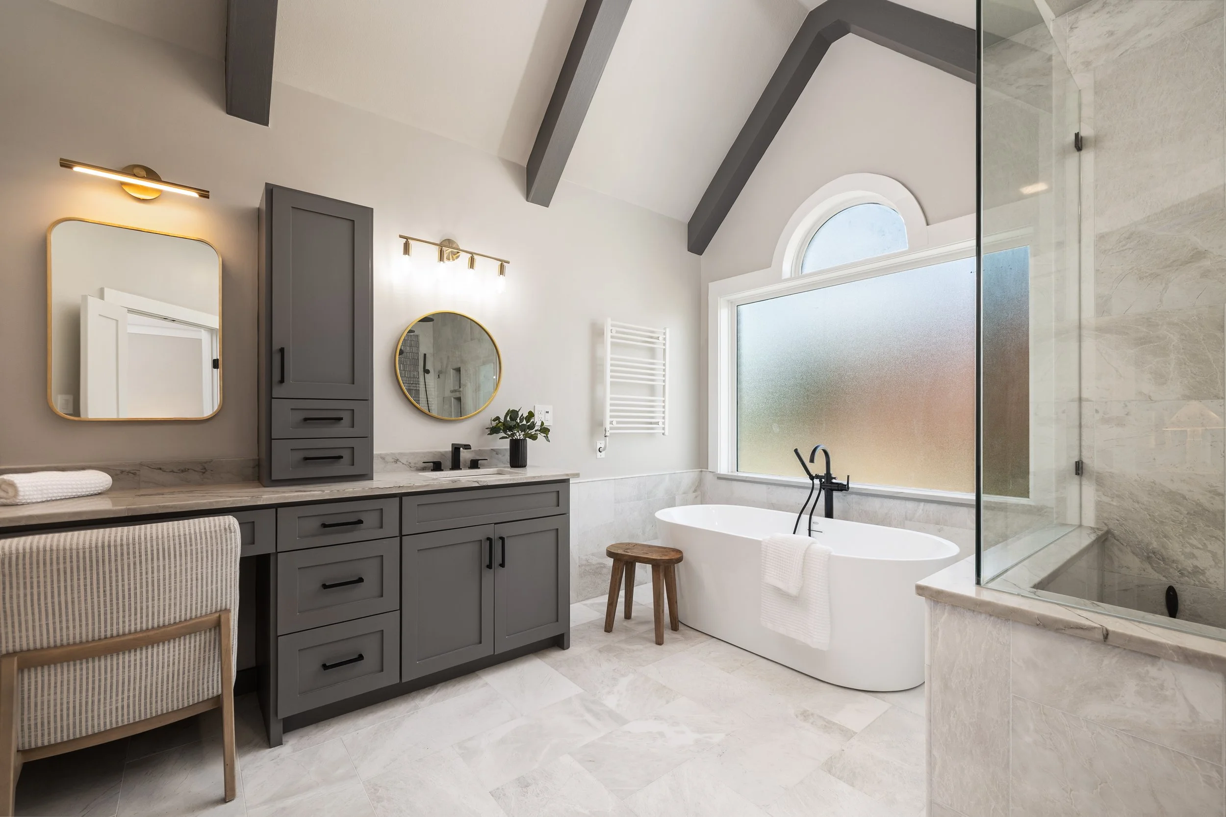 Modern bathroom with a white bathtub, gray cabinets, a black faucet, a large frosted window, and beige tile flooring.