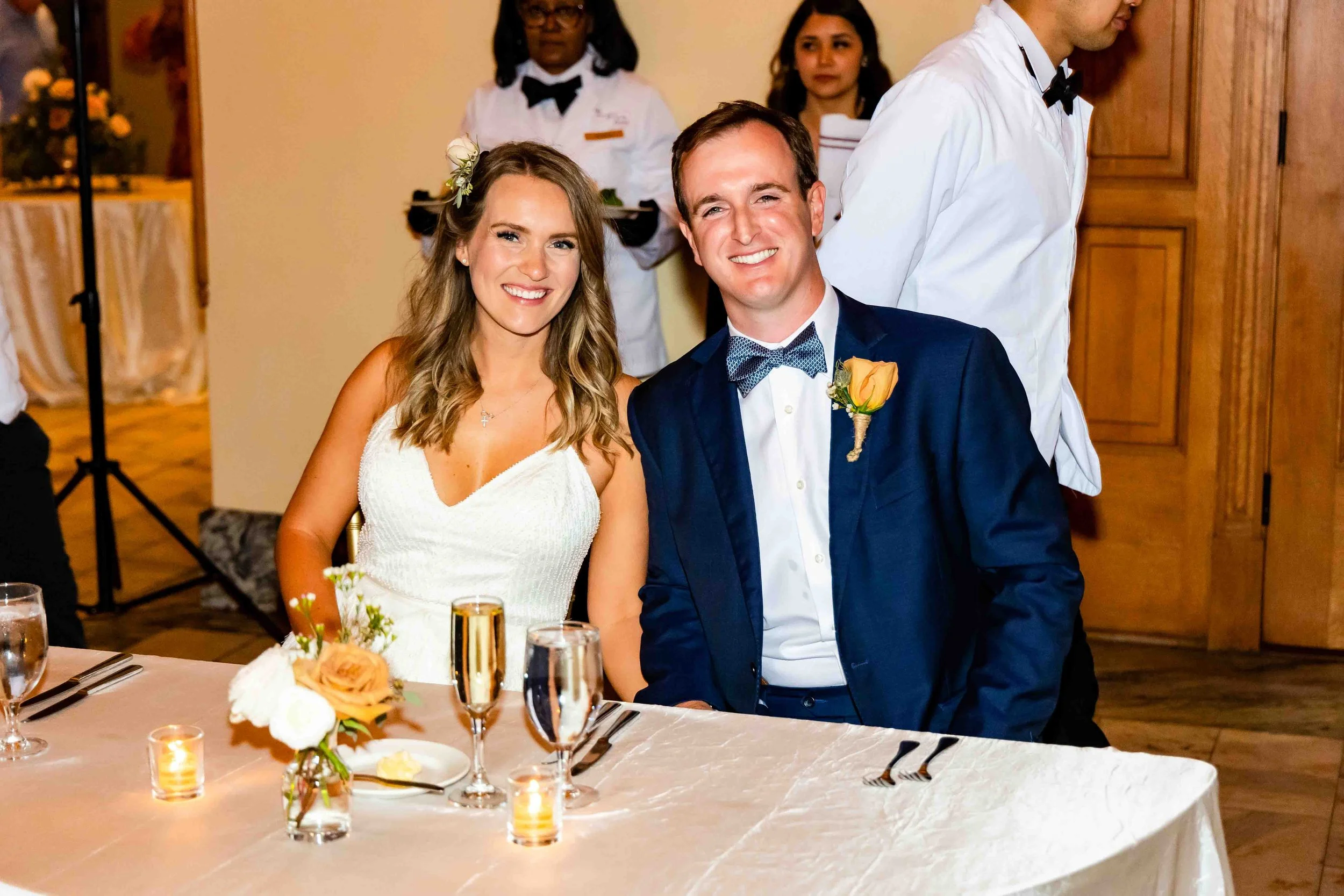 A couple at a wedding reception, sitting at a table with floral centerpieces and candles, with waitstaff in the background.
