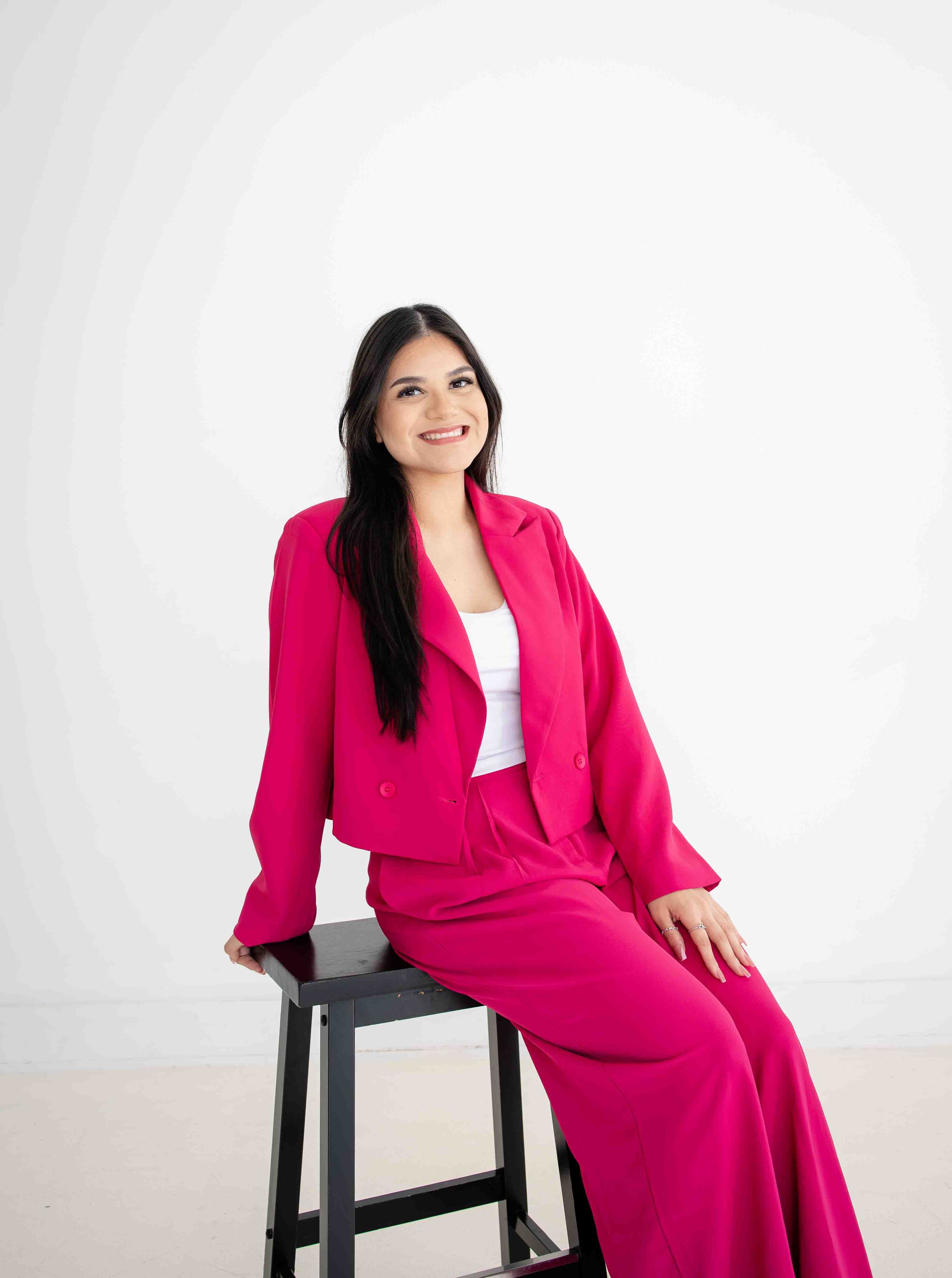 Woman in pink suit smiling, sitting on stool against white background.