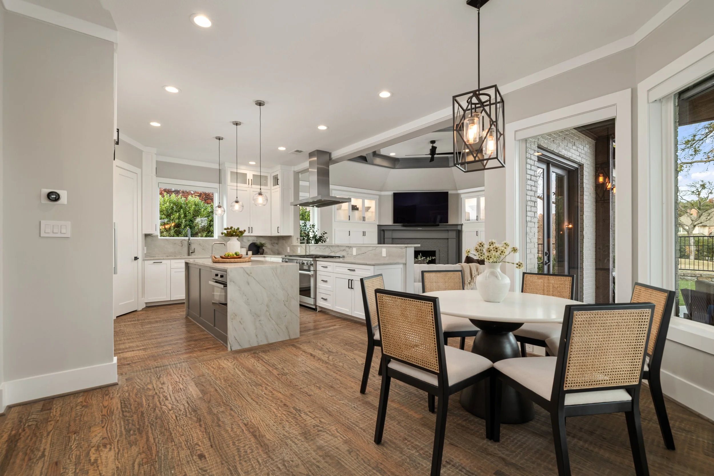 Modern kitchen and dining area with white cabinets, marble island, wooden floor, and large windows.