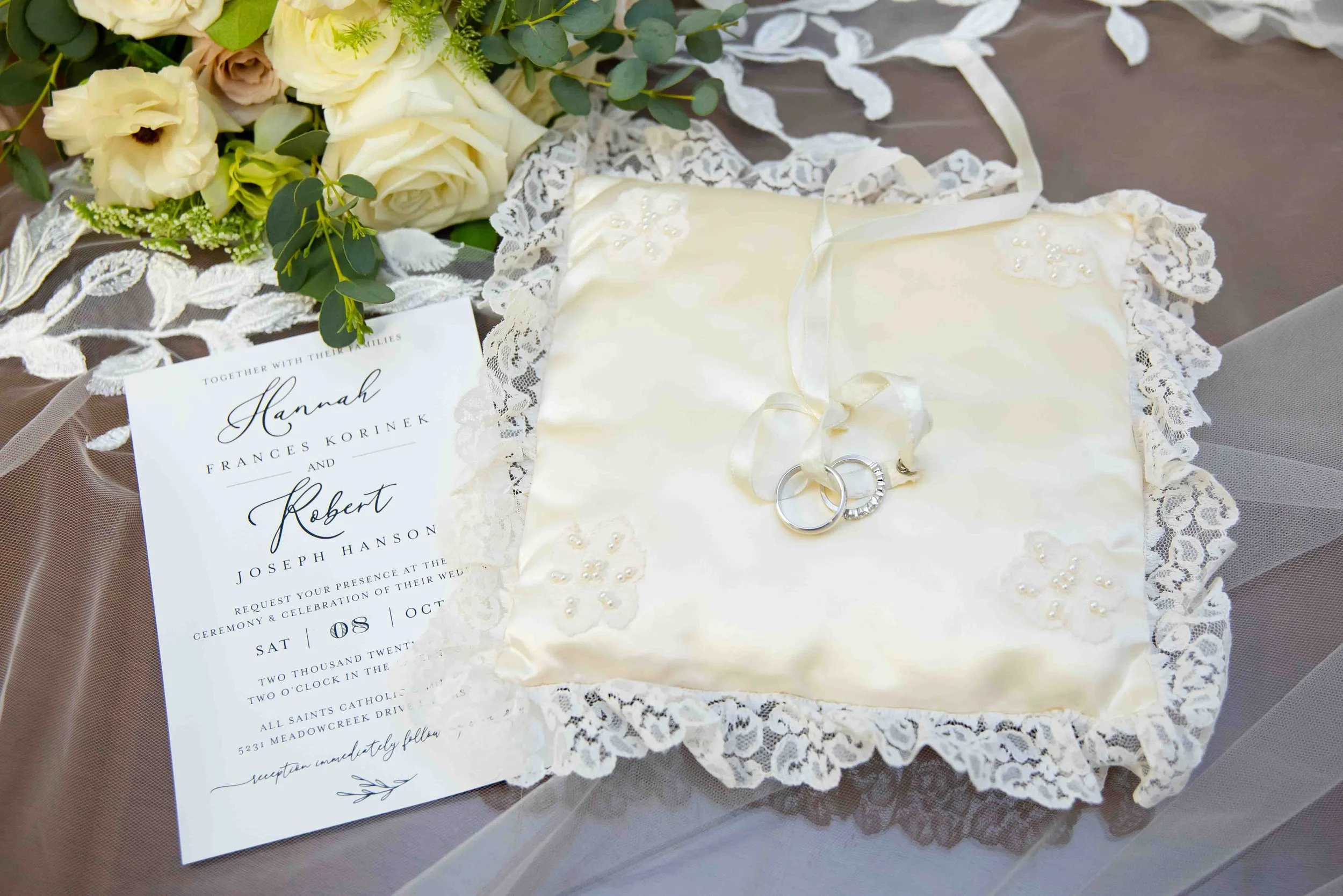 Wedding invitation, bouquet of white and cream flowers, and wedding rings on a satin pillow with lace edges.