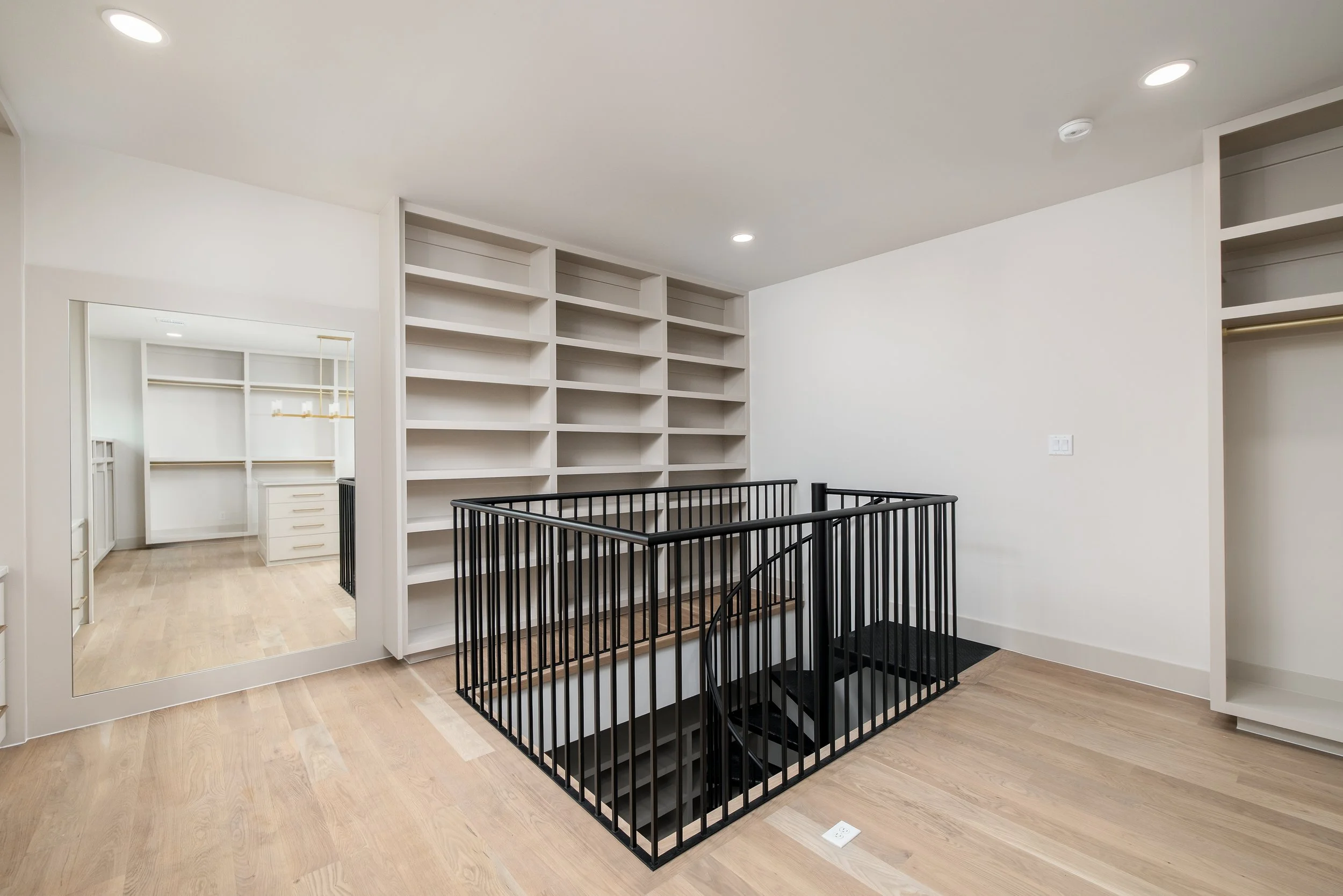 An empty room with built-in white bookshelves, a black metal railing around a staircase, a large mirror, and open closet space.