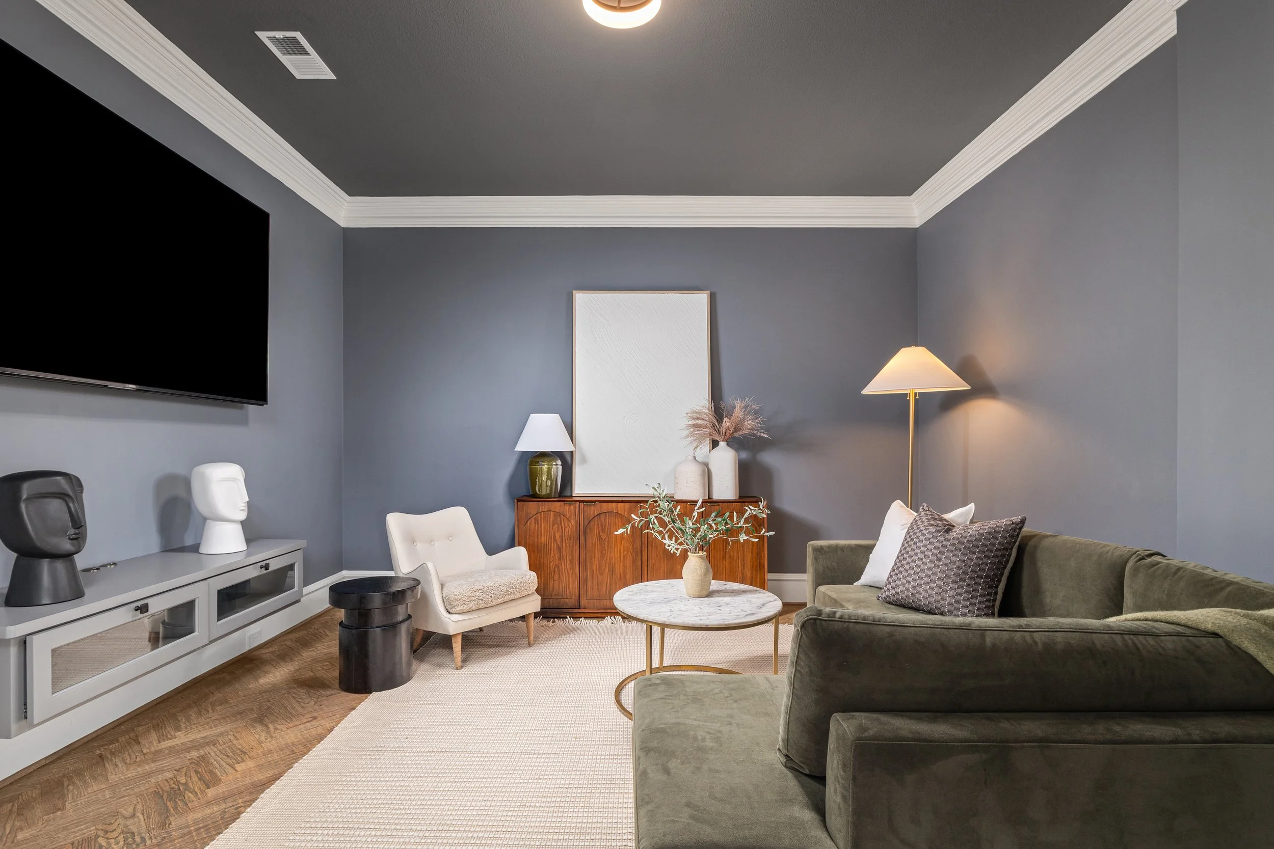 Living room with dark green sofa, white armchair, wooden sideboard with vases and lamp, wall-mounted TV, and farmhouse decor.