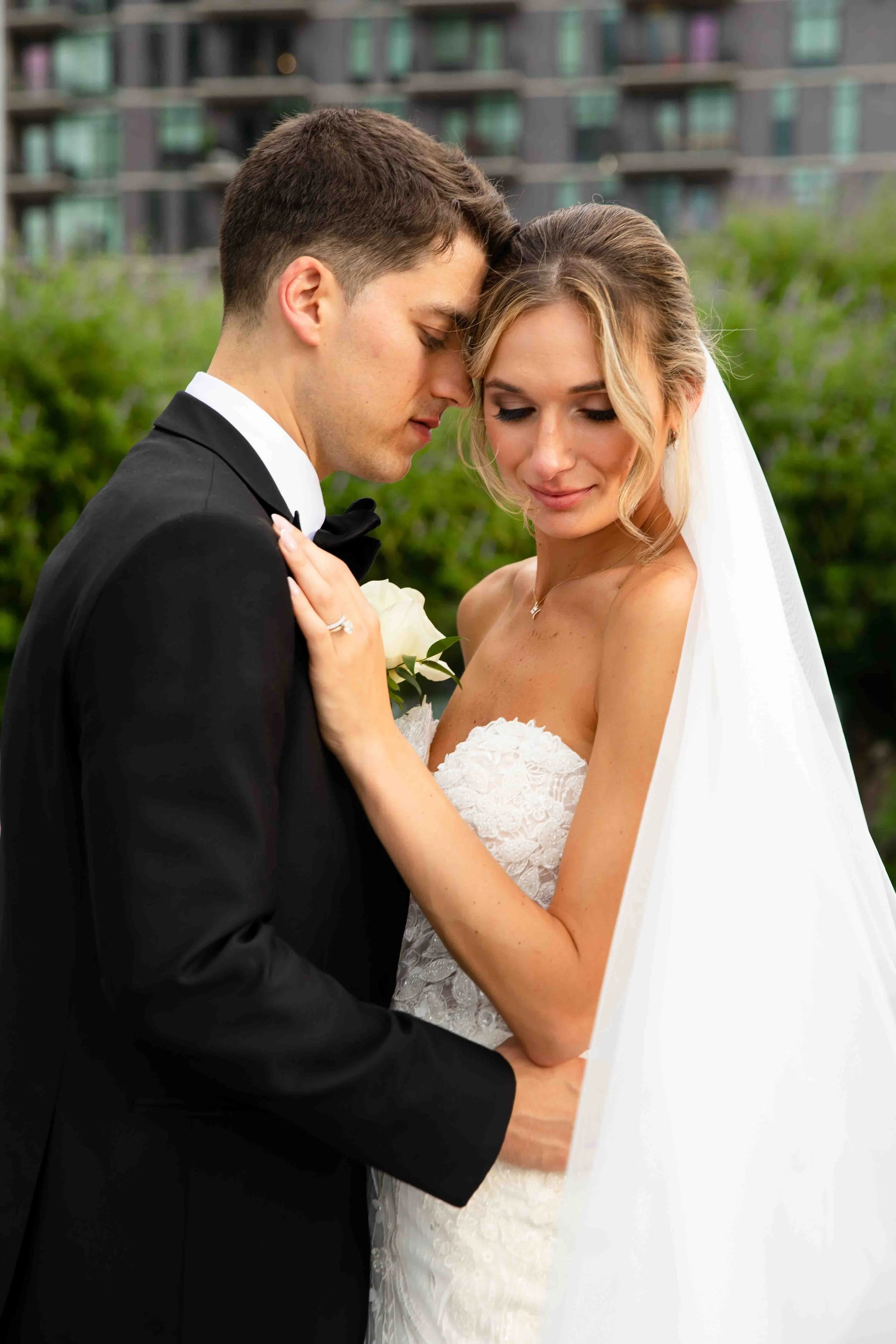 A bride and groom sharing an intimate moment outdoors, with the groom in a tuxedo and the bride in a strapless wedding dress and veil.