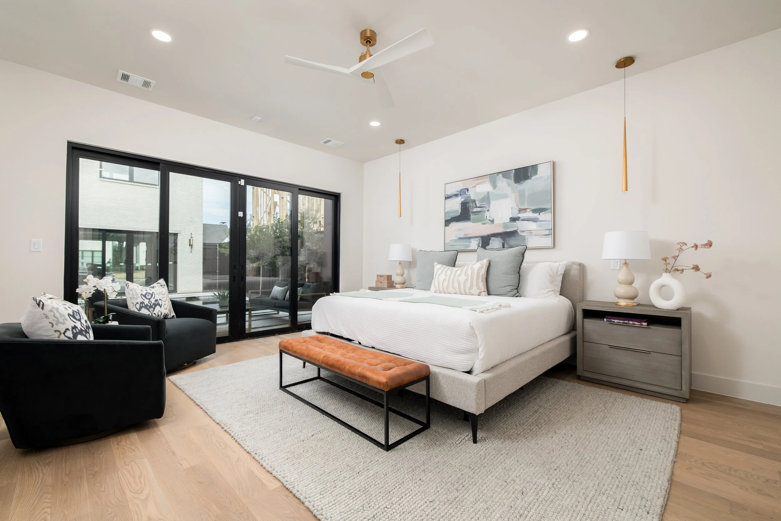 Modern bedroom with white walls, beige upholstered bed, abstract wall art, two matching nightstands with lamps, black armchair with patterned pillows, glass sliding door leading to outdoor patio, light wood flooring, ceiling fan, beige area rug, oran