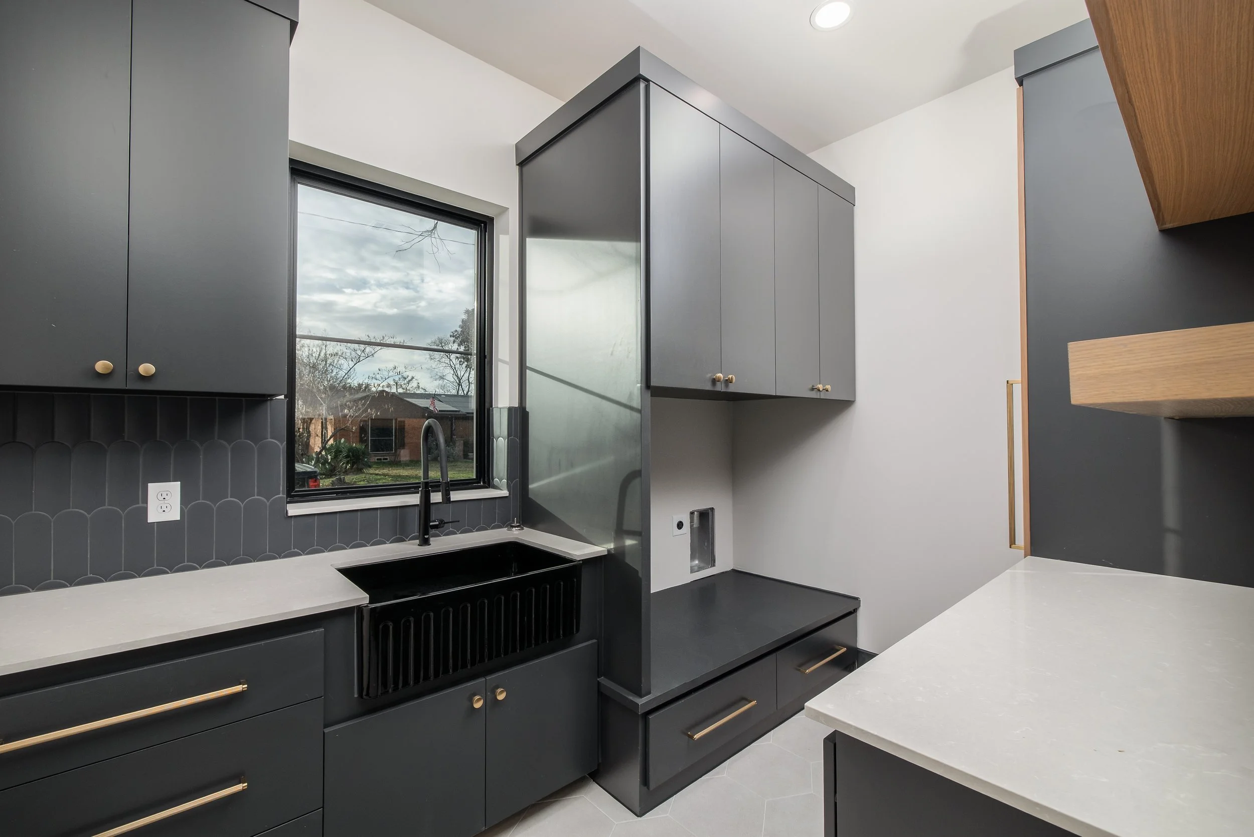 Modern kitchen with gray cabinets, black sink, white countertops, and a large window showing a suburban neighborhood outside.