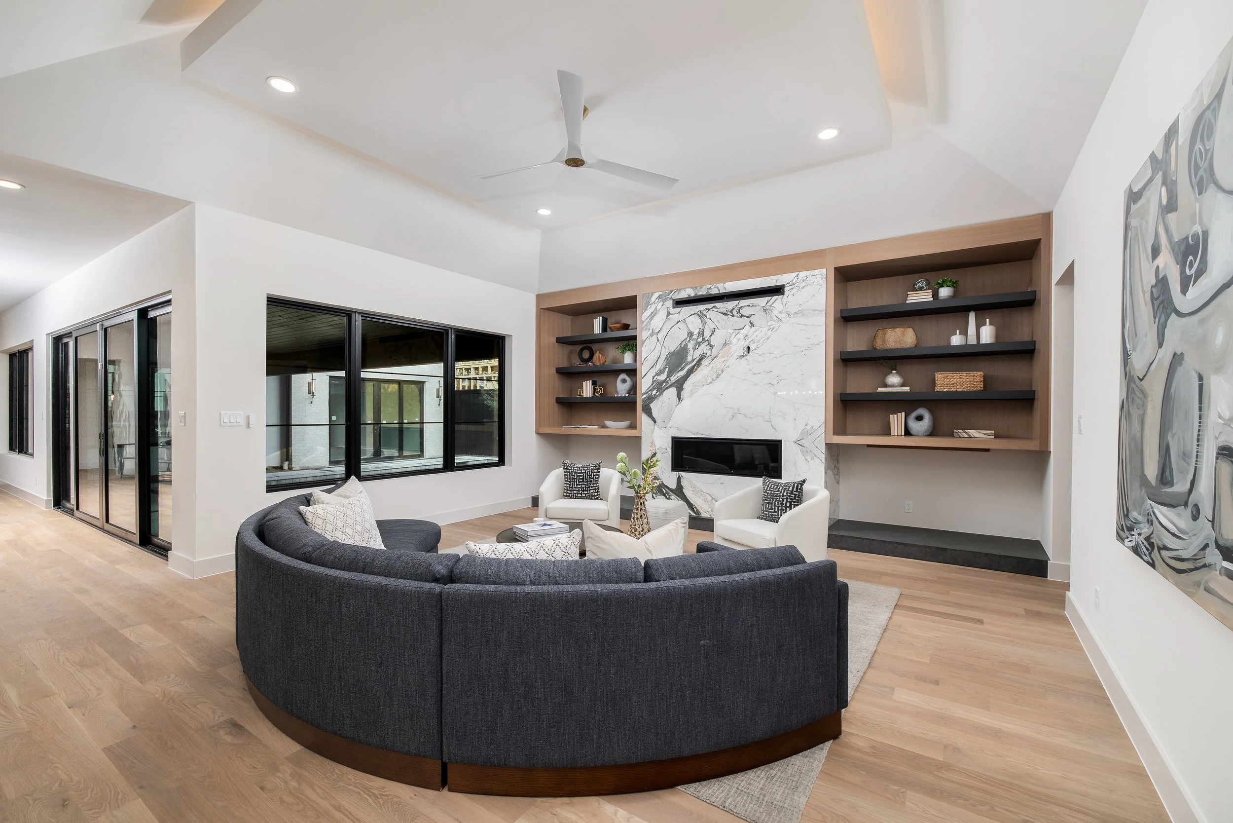 Modern living room with a curved gray sofa, white armchairs, and decorative pillows. Features a marble fireplace with built-in shelves, large windows, and a ceiling fan.