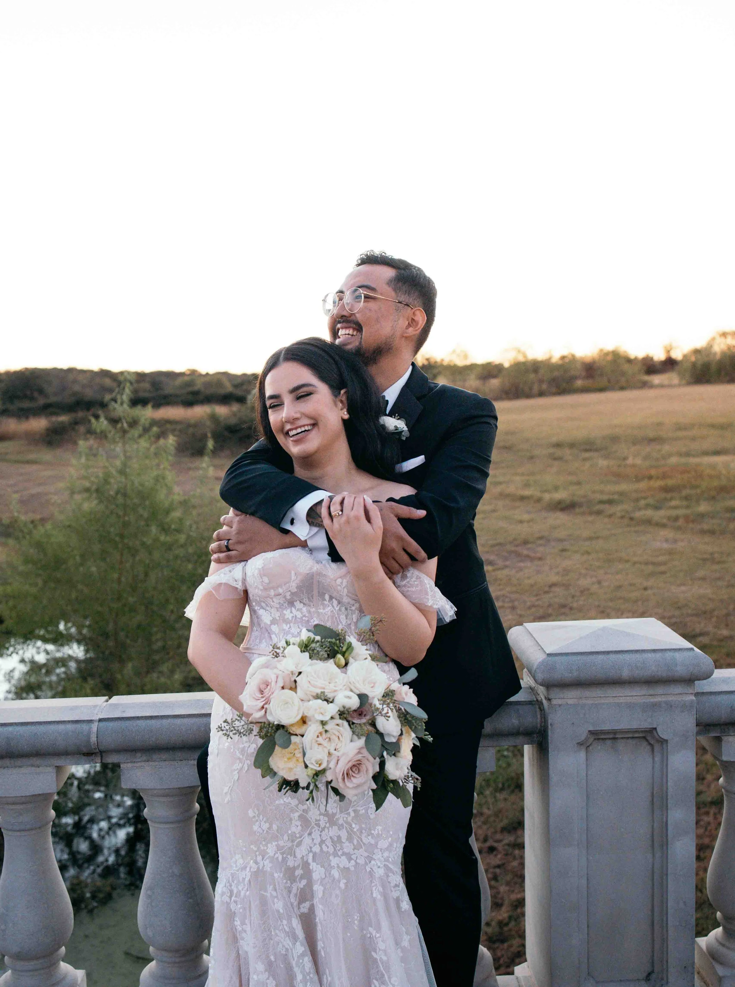 A bride and groom sharing a joyful moment outdoors on a bridge, with the bride holding a bouquet of flowers and the groom hugging her from behind, during sunset.