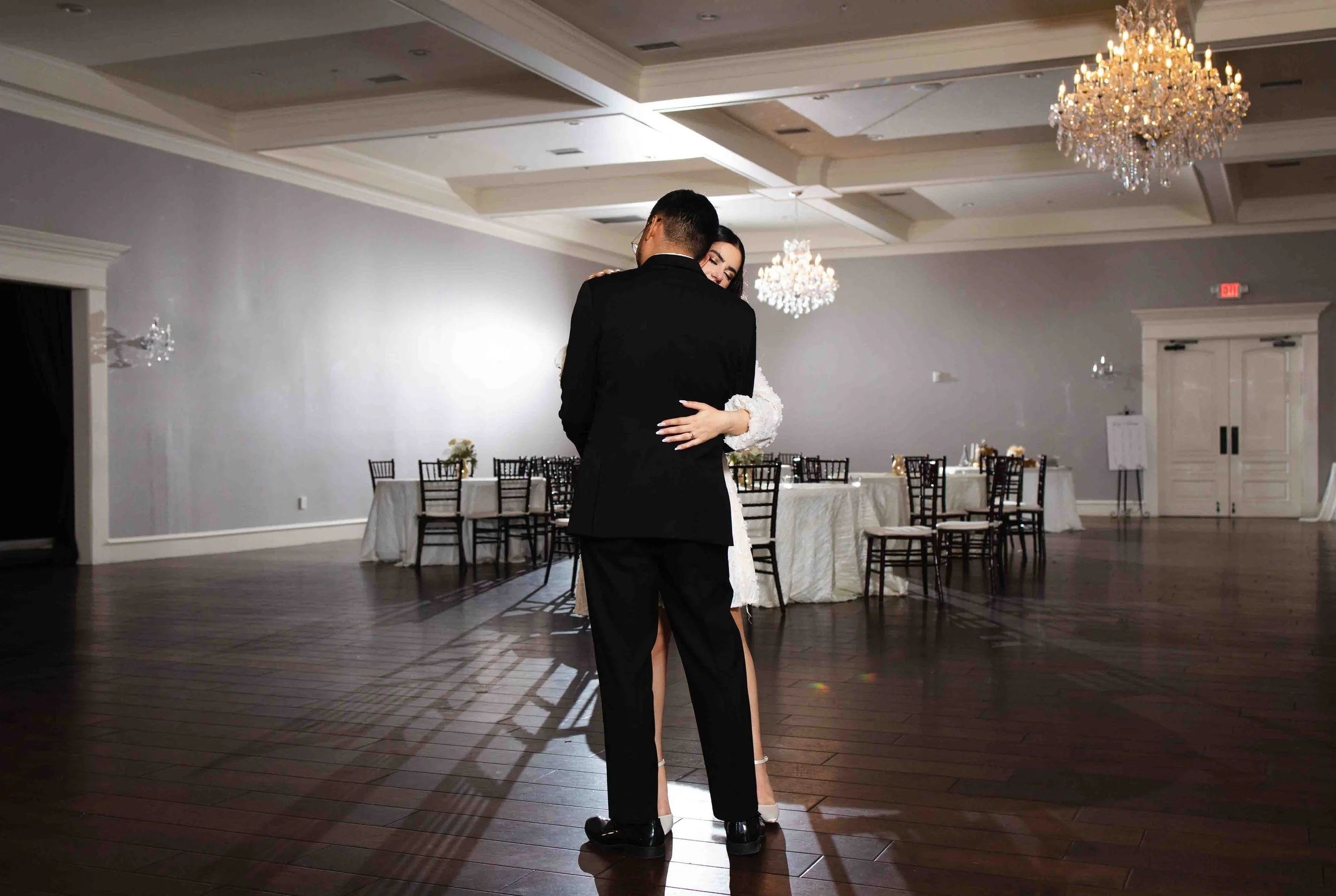 A couple dancing in a ballroom with dark wooden floors, chandelier lighting, and decorated tables in the background.