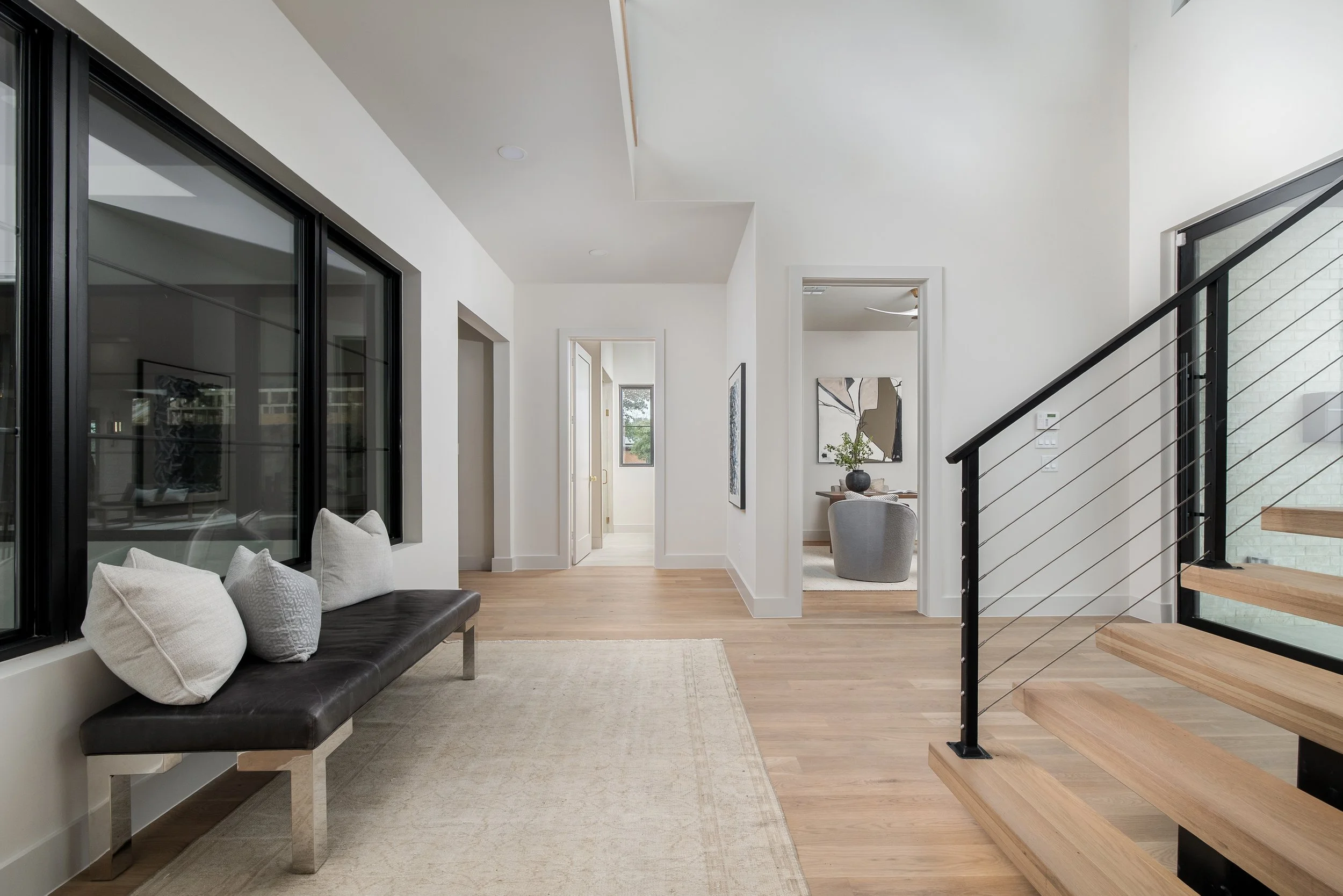 Modern living space with white walls, hardwood floors, large black-framed windows, a black bench with beige pillows, a beige rug, and a staircase with wooden steps and metal railing.