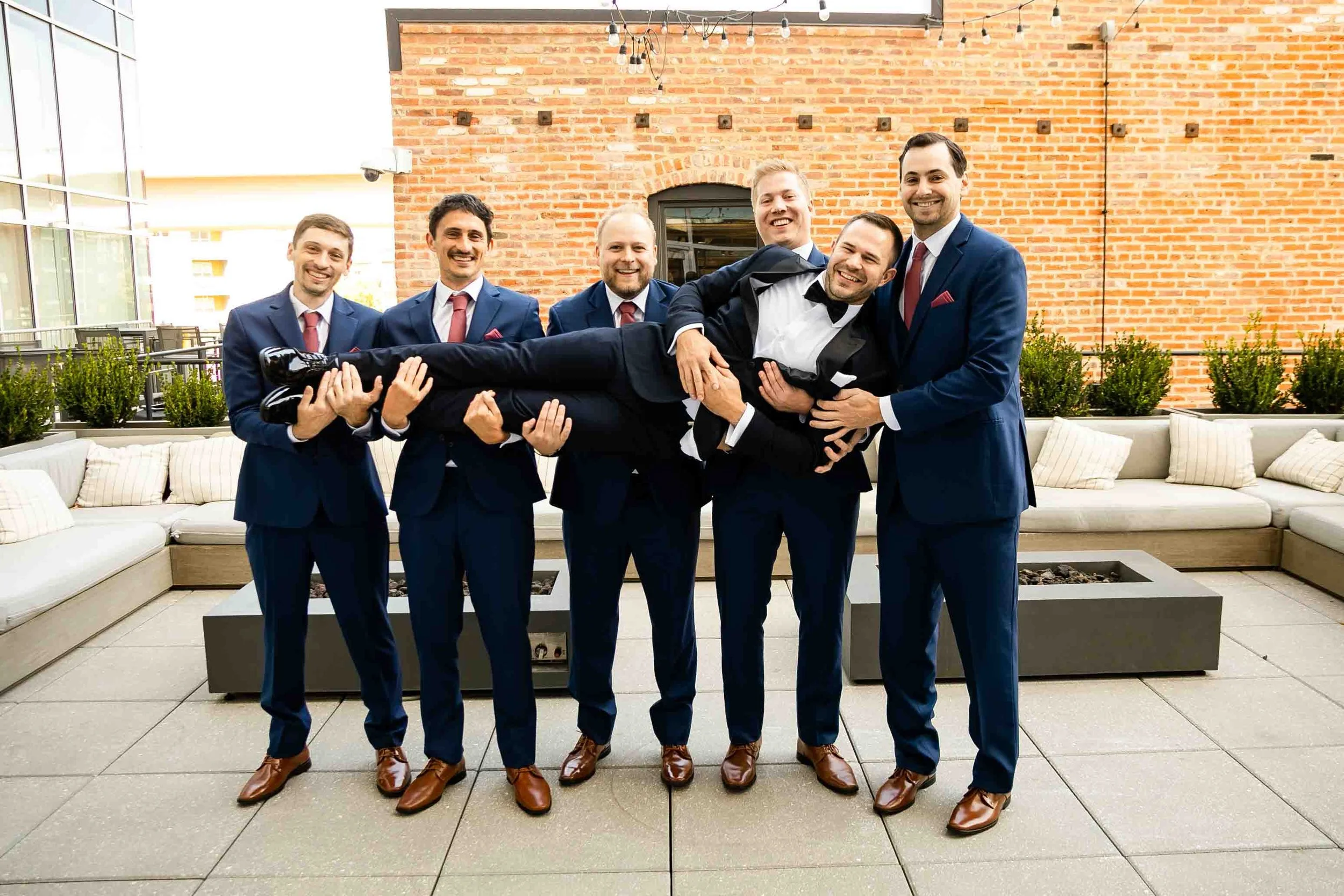 A group of seven men in formal suits, with five men holding a man in a tuxedo horizontally, smiling and laughing on an outdoor patio with brick and glass walls and string lights overhead.