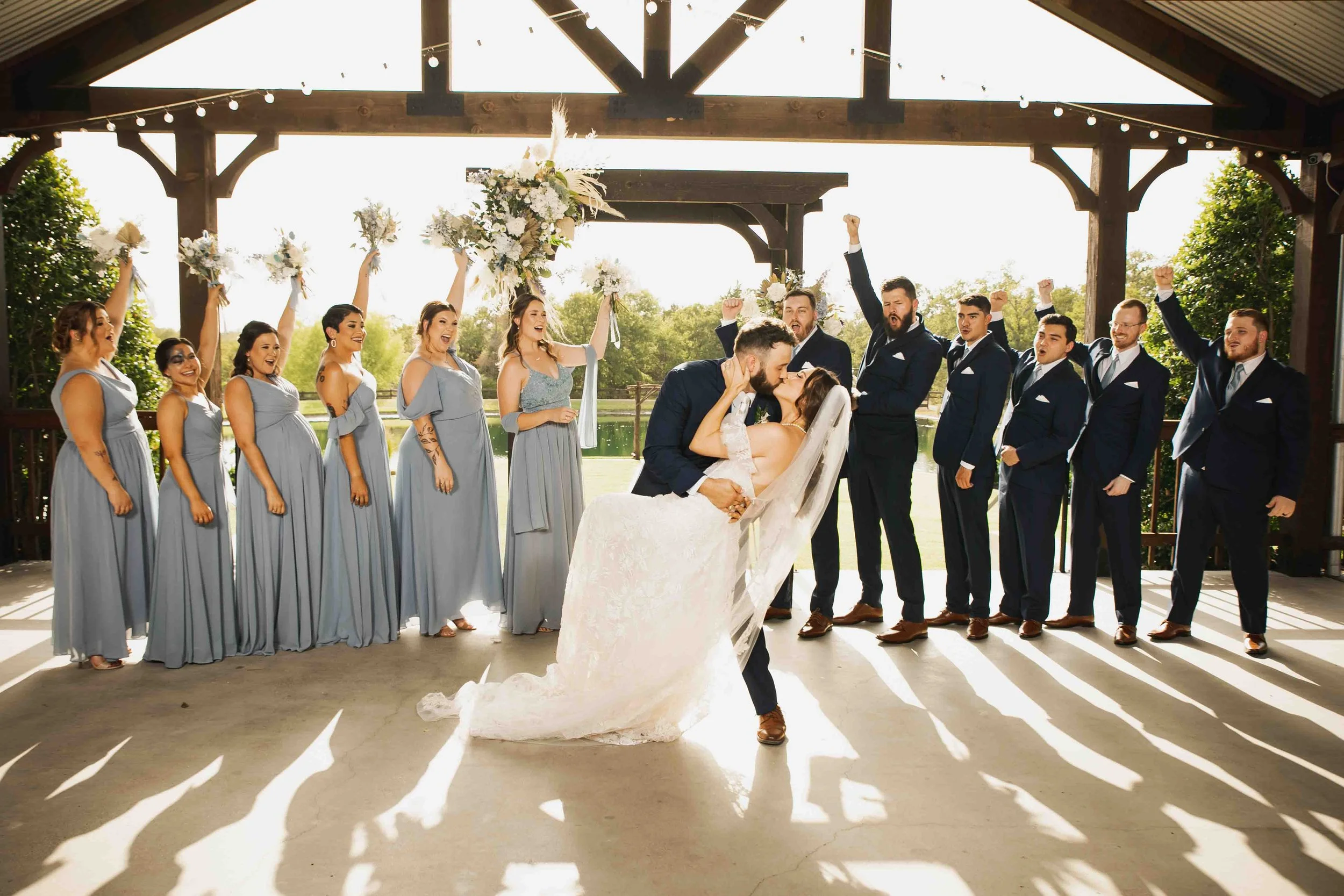 A wedding celebration with a bride and groom kissing, surrounded by bridesmaids and groomsmen in formal attire, under a pavilion with string lights and outdoor scenery
