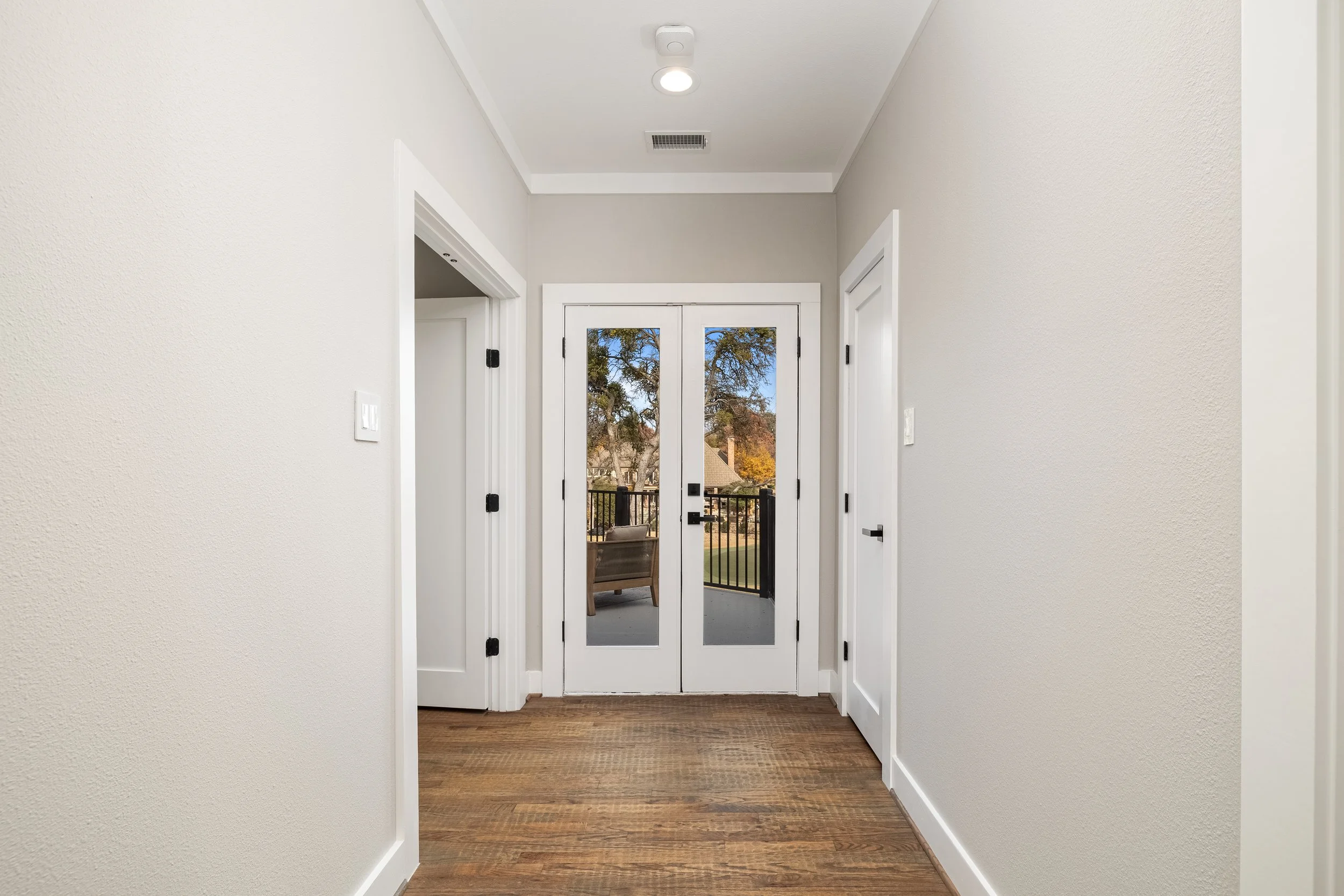 Interior hallway with double glass doors leading to a balcony, wooden floor, white walls, and white door frames.