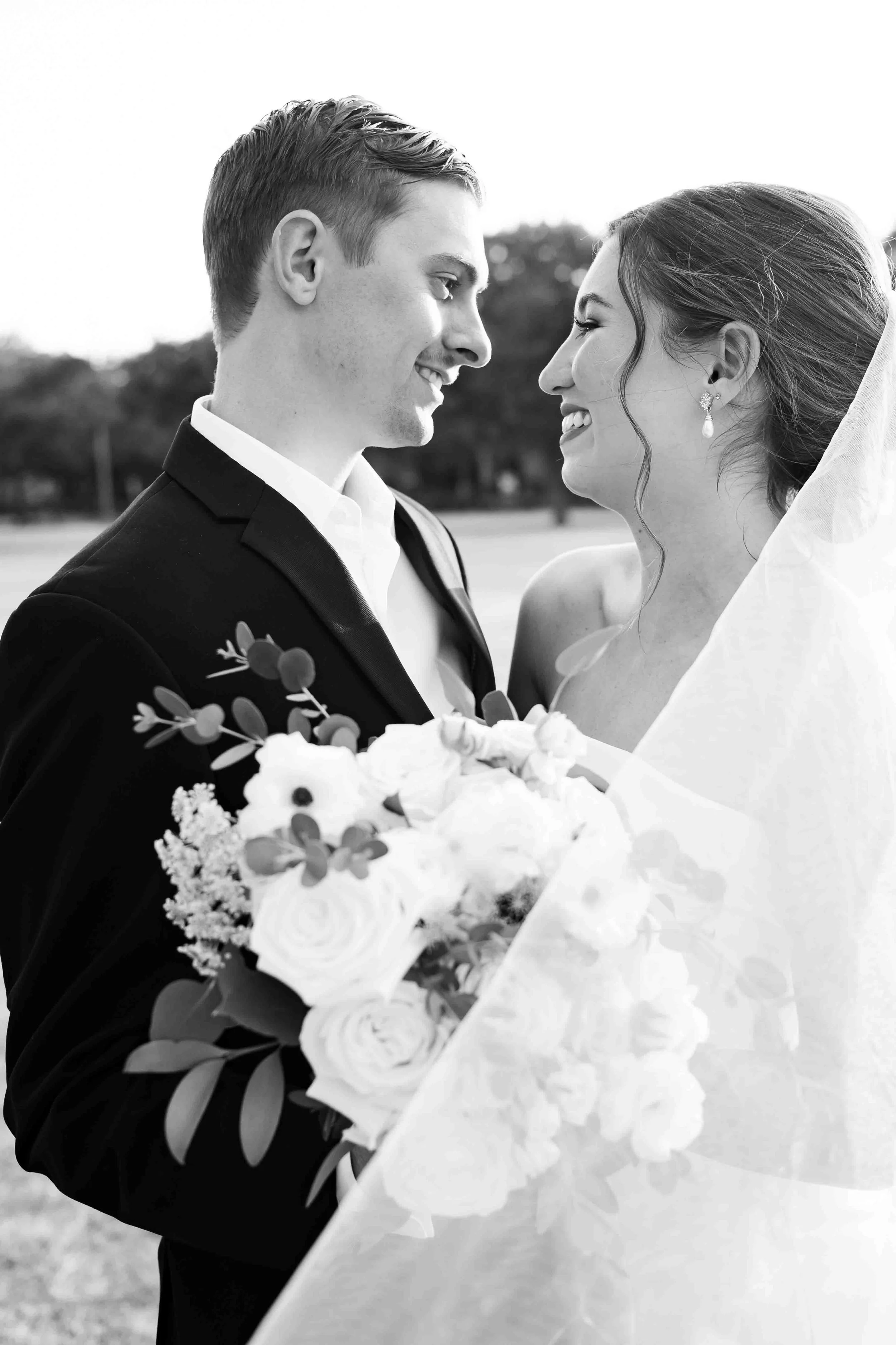Black and white photo of a bride and groom smiling at each other outdoors, with the groom holding a bouquet of flowers.
