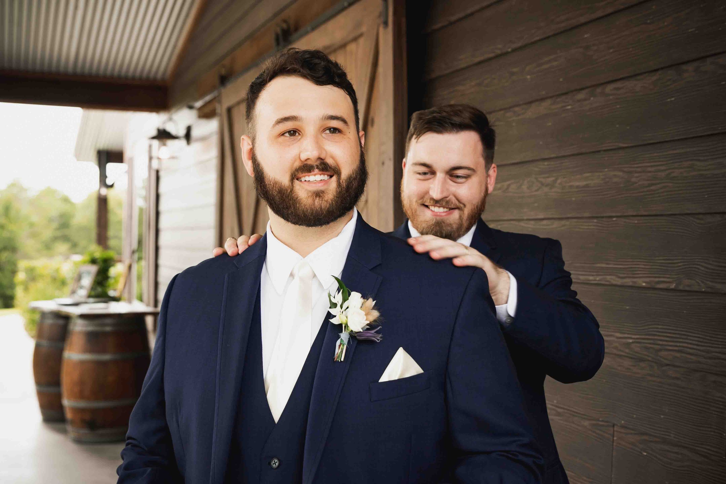Two men in suits, smiling, one adjusting the other's shoulder, outdoors near a wooden building, during a wedding or special event.