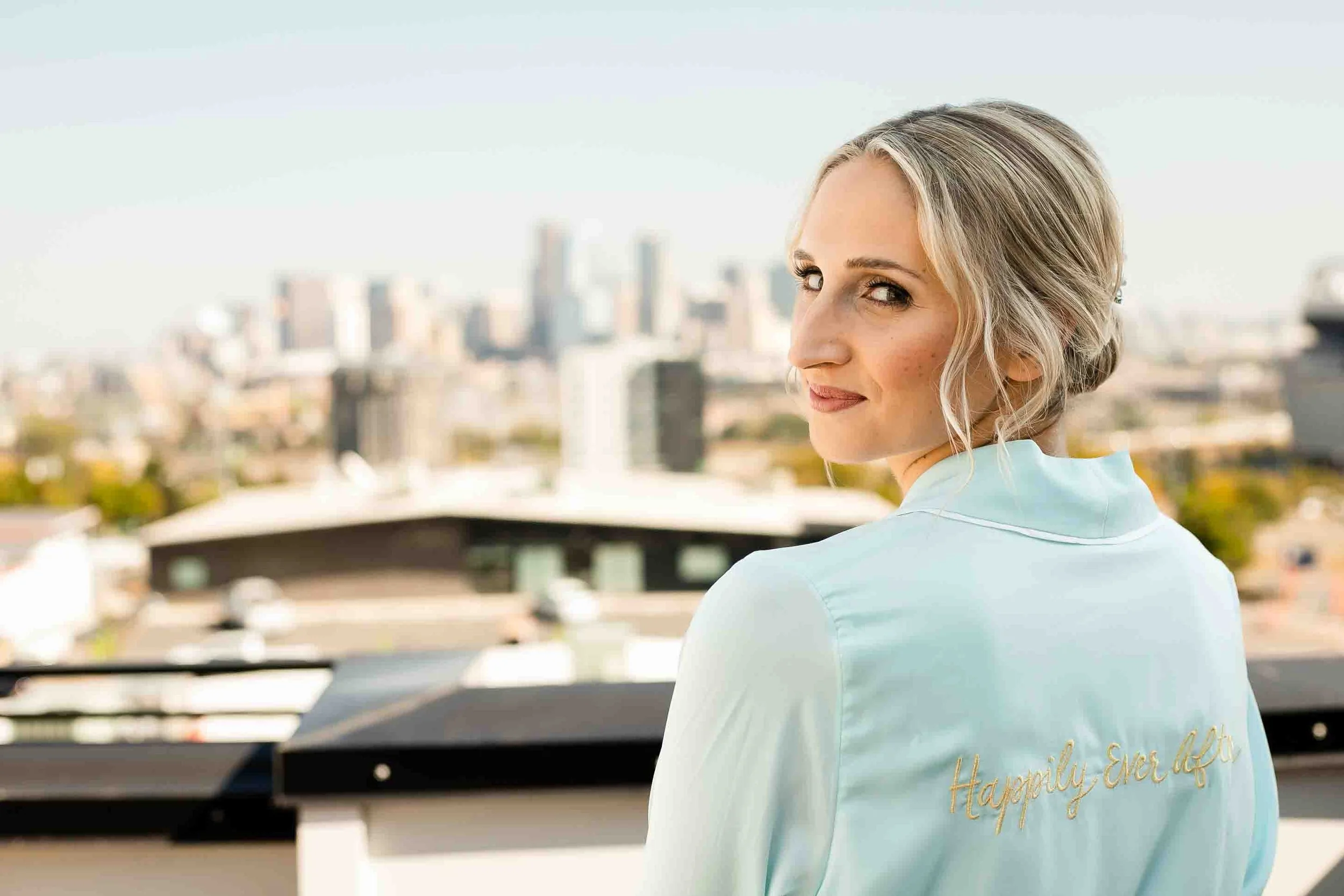A woman with blonde hair styled in waves, wearing a satin robe with the words 'Happily Ever After' embroidered on the back, looking over her shoulder at a city skyline.