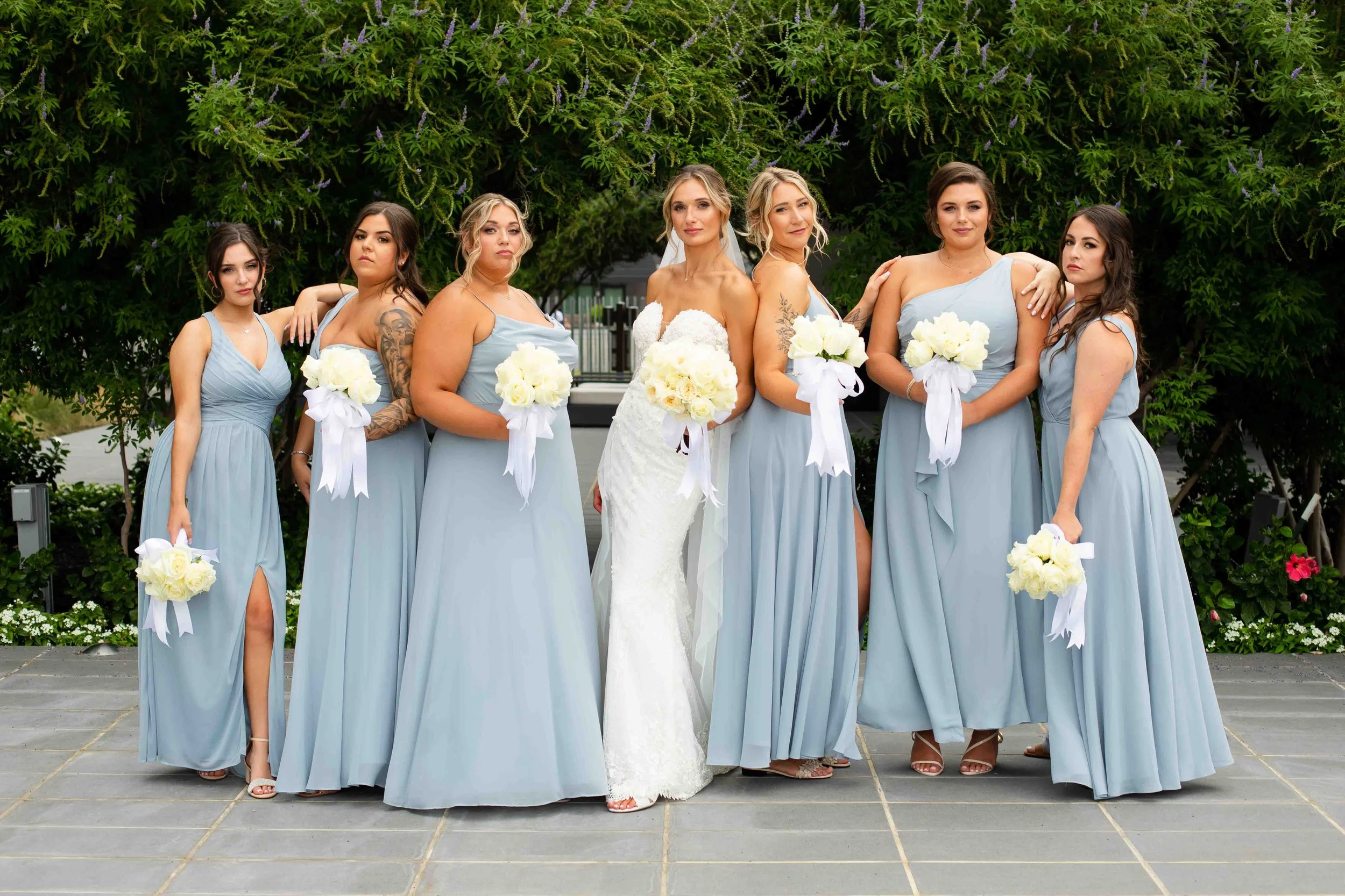 Bride in a white wedding dress with six bridesmaids in light blue dresses holding white bouquets, standing outdoors in front of green trees and bushes.