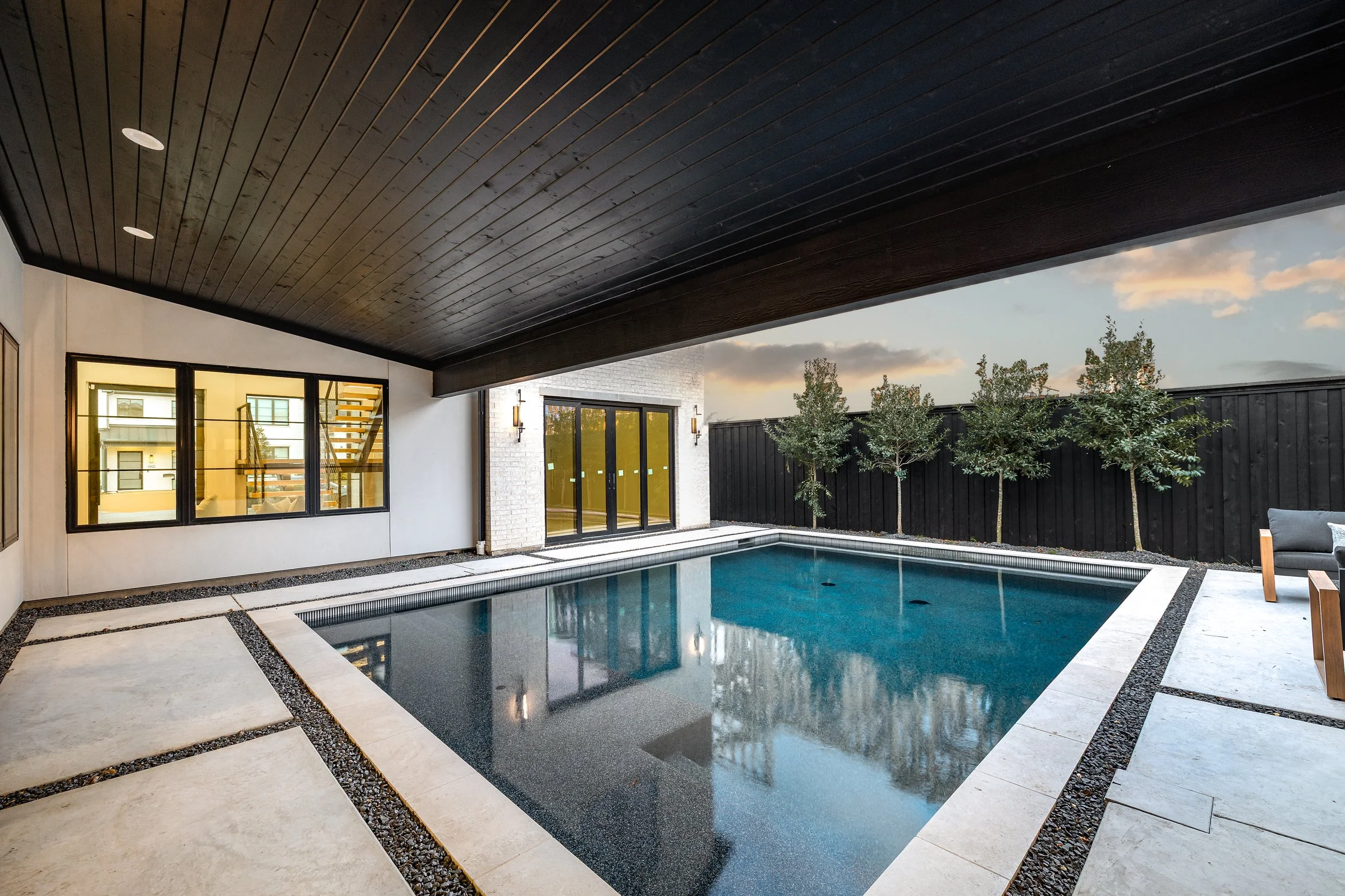 Modern backyard with a rectangular swimming pool, surrounded by beige stone tiles, black wooden fence, and four small trees, with a covered patio area and a house with large windows and a sliding glass door.