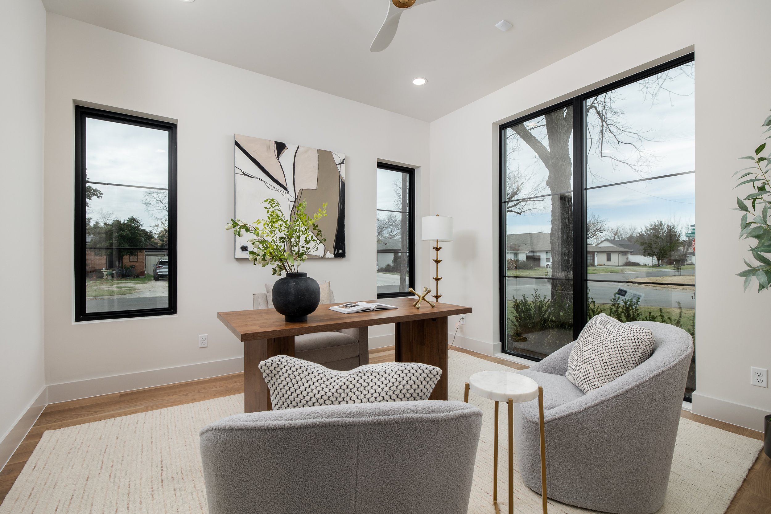Modern living room with large windows, contemporary chairs, a wooden desk, wall art, and a potted plant.
