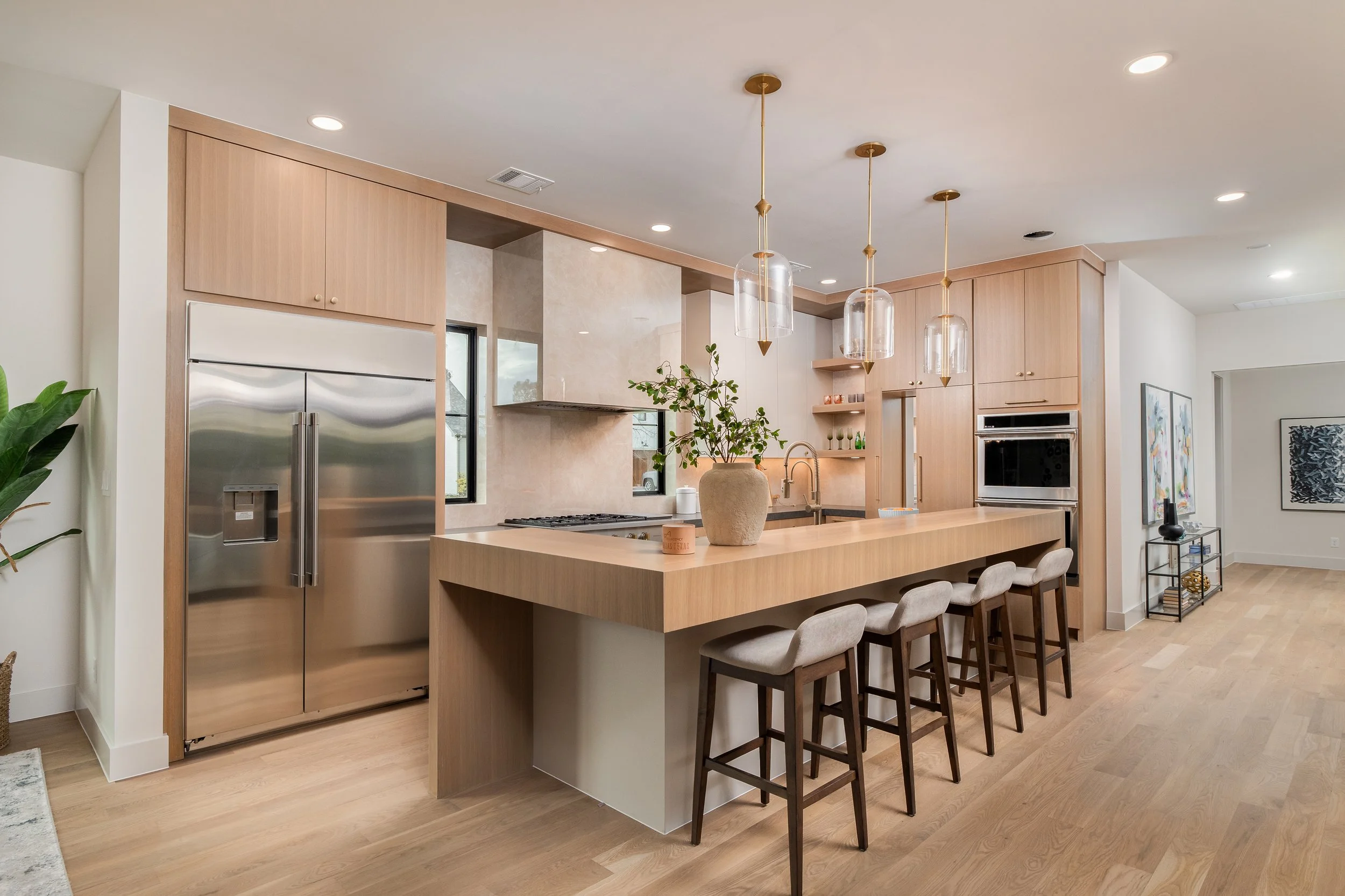 Modern kitchen with light wood cabinets, a large island with bar stools, stainless steel refrigerator, built-in oven, and pendant lights, with minimal decor and hardwood flooring.