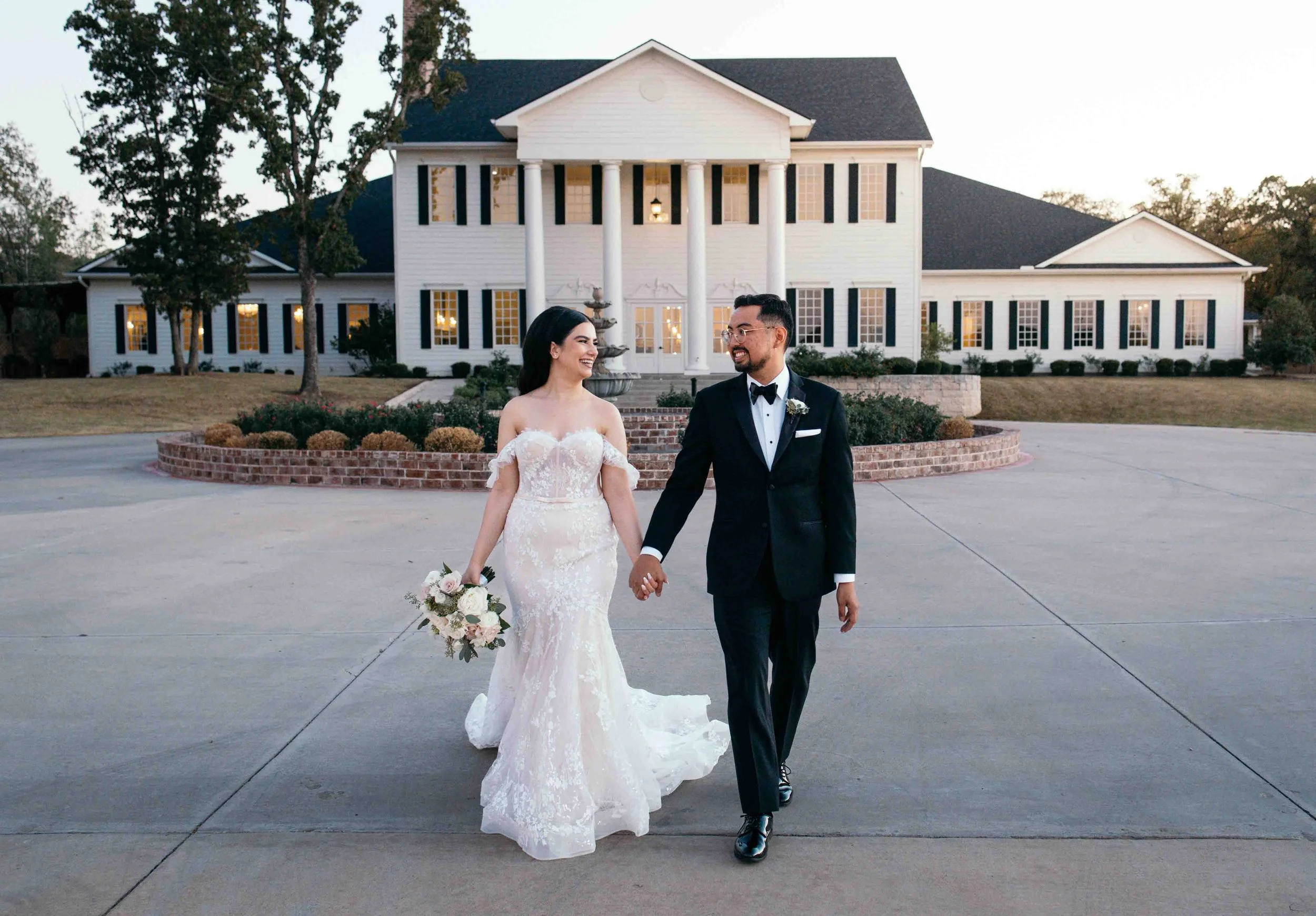 A newlywed couple walking hand in hand outside a large white house with black shutters during sunset.