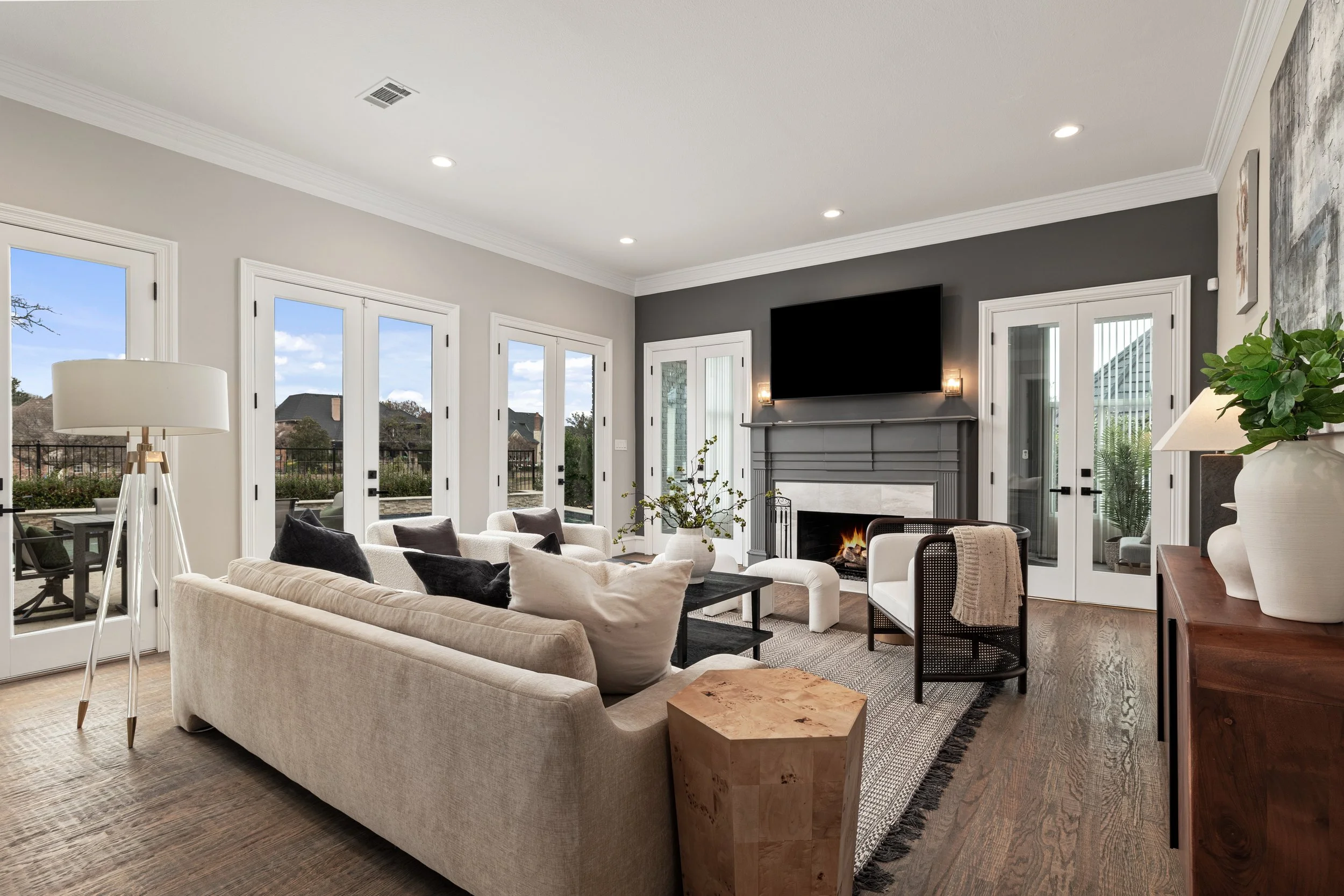 Living room with beige sofa, black and white pillows, armchair with a throw blanket, fireplace with TV above, large windows and glass doors, wooden floor, and potted plants.