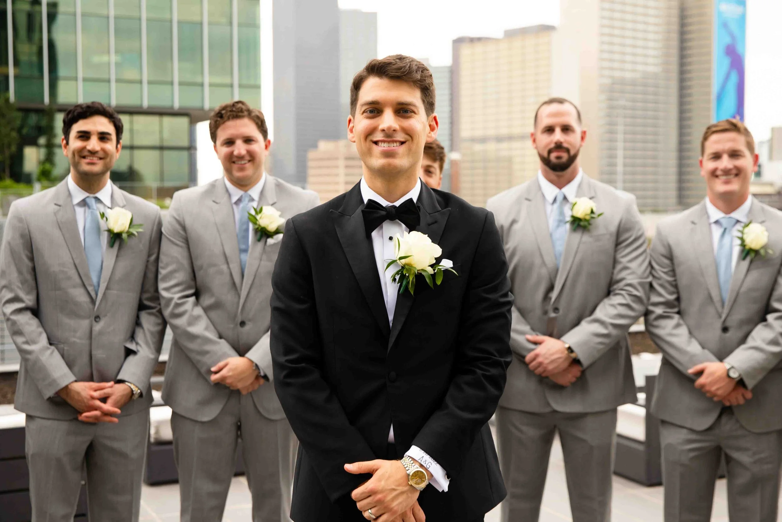 A groom in a black tuxedo and bow tie, smiling, standing in front of five groomsmen in light gray suits with white shirts and light blue ties, all with white boutonnieres, on a rooftop with a city skyline in the background.