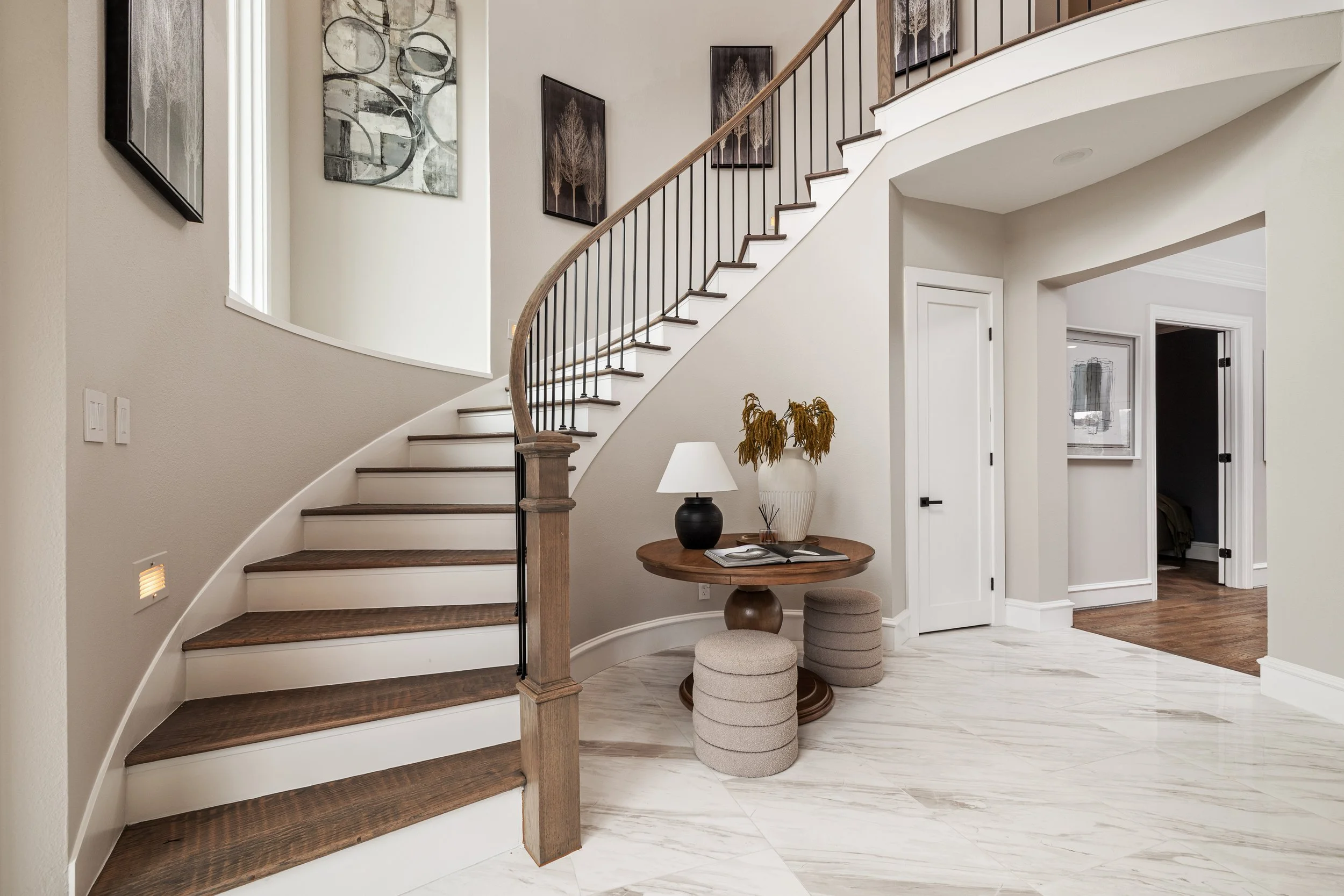 Modern staircase with dark wooden steps and black metal railing in a bright entryway, with framed artwork on the walls, a small round table with a lamp and a vase with dried plants, and marble flooring.