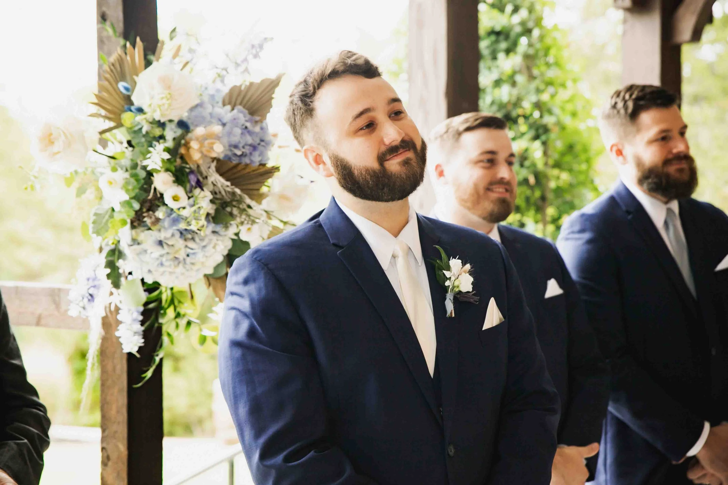 Groom with beard in navy suit standing beside groomsmen at wedding ceremony, floral arrangement in background