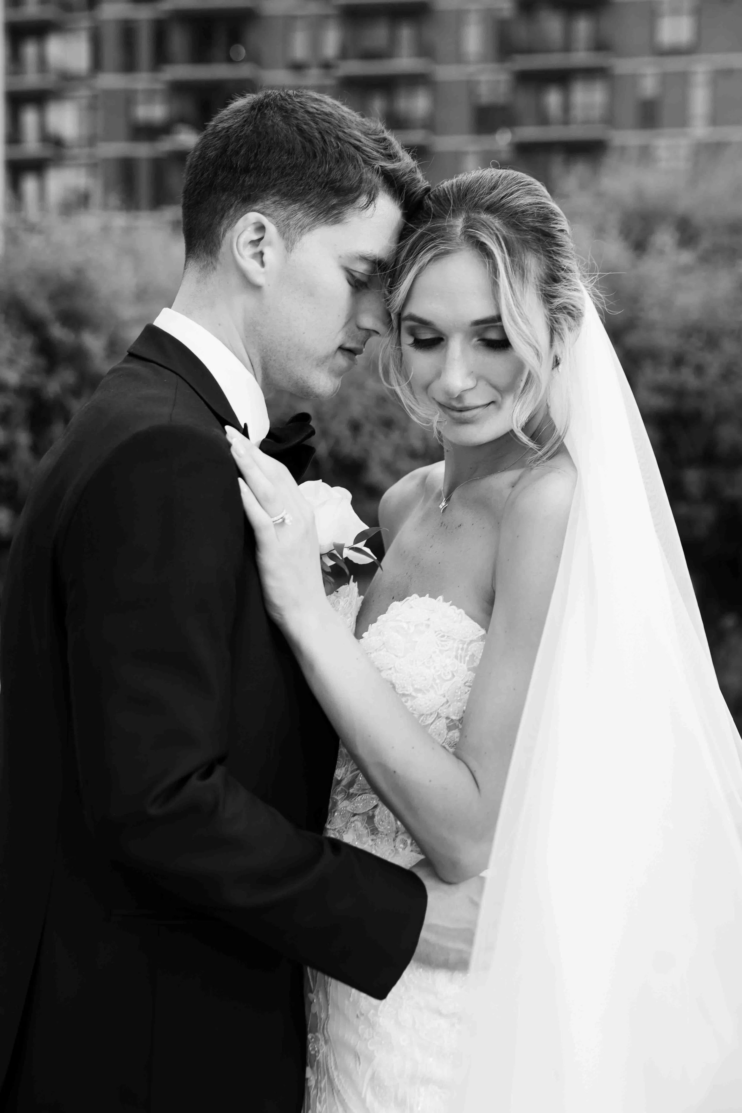 Black and white photo of a bride and groom on their wedding day, embracing each other with their foreheads touching, outdoors with a blurred background of buildings and trees.