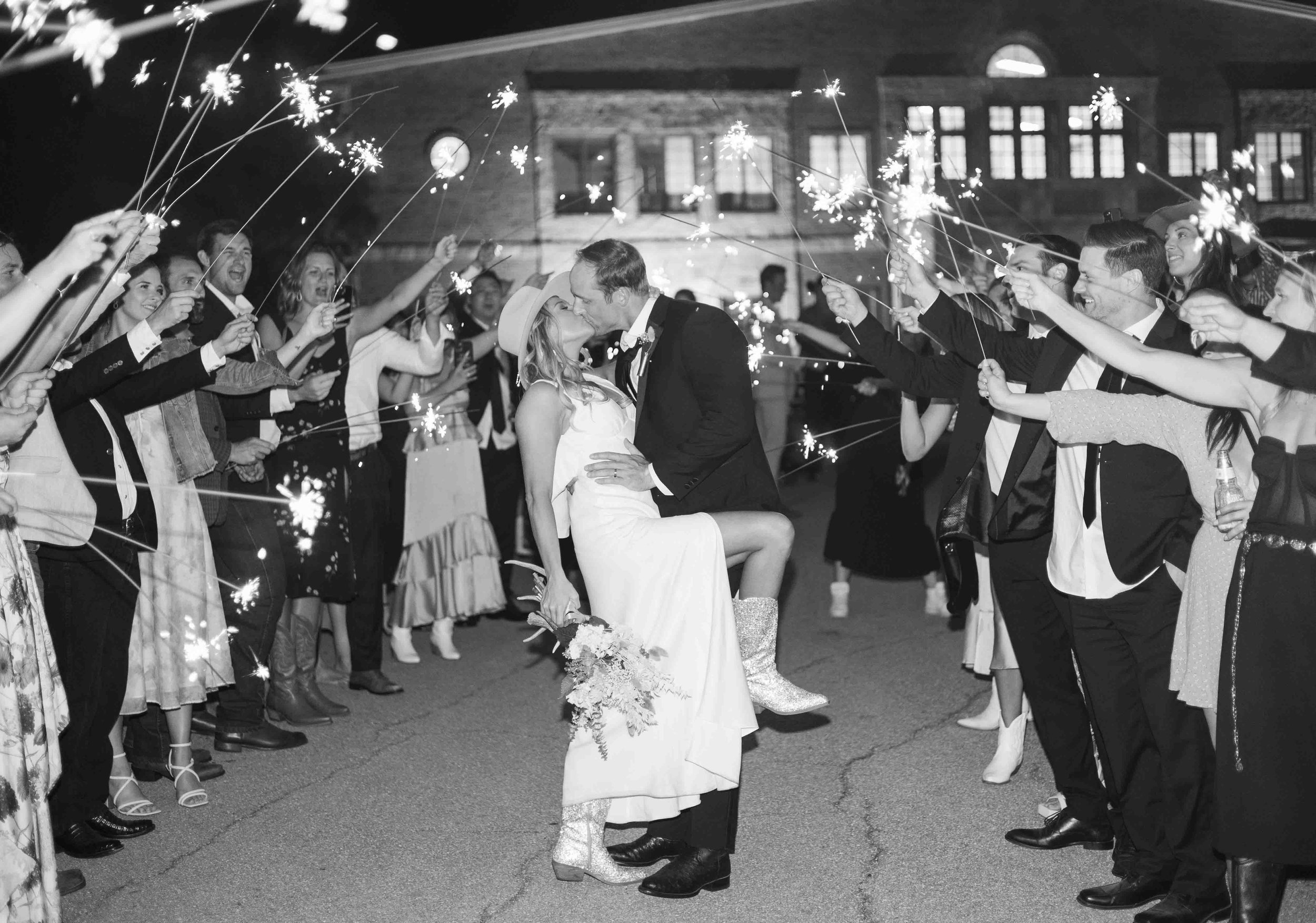 A couple dressed in wedding attire sharing a kiss while being lifted, surrounded by friends holding sparklers at night outside a building.