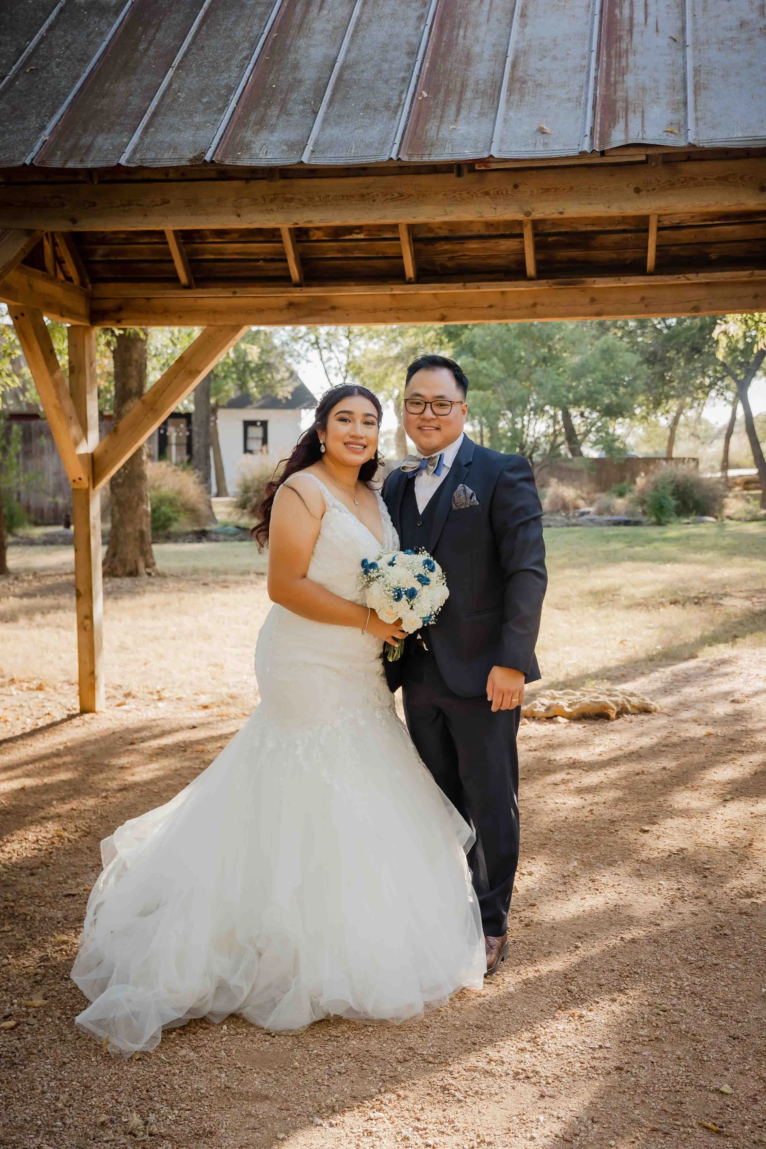 A bride and groom standing together under a wooden shelter outdoors, smiling at the camera. The bride is wearing a white wedding gown and holding a bouquet of white roses with blue accents. The groom is dressed in a navy suit with a bow tie and glass
