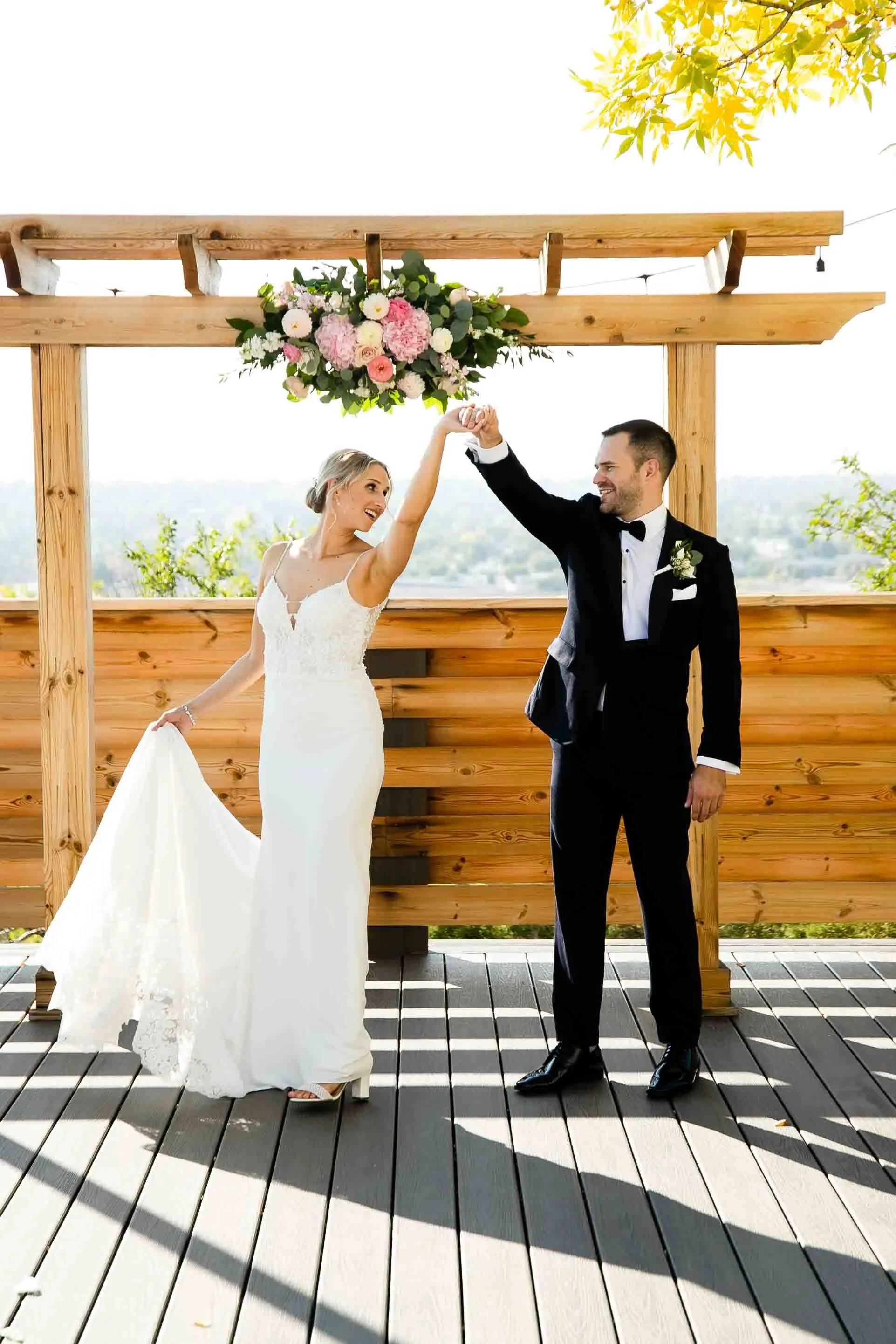 Bride and groom dancing at their wedding ceremony outdoors, with a floral arrangement hanging above them and a wooden backdrop.