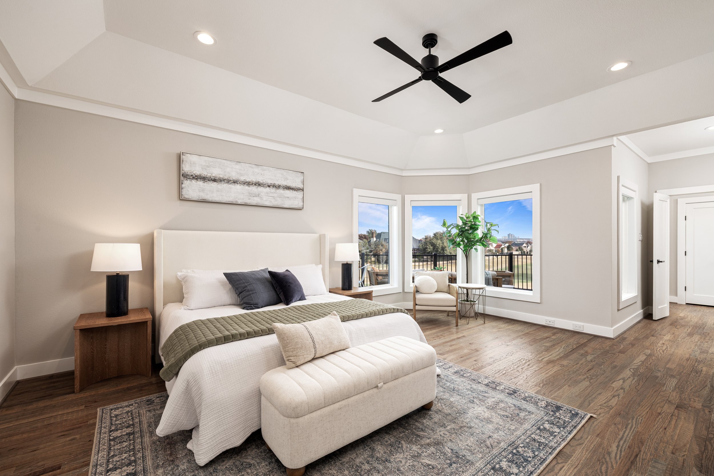 Bright bedroom with large windows, white bed with gray and black pillows, wooden nightstands with lamps, beige bench at foot of bed, area rug, small chair, small tree, wooden flooring, ceiling fan, and recessed lighting.