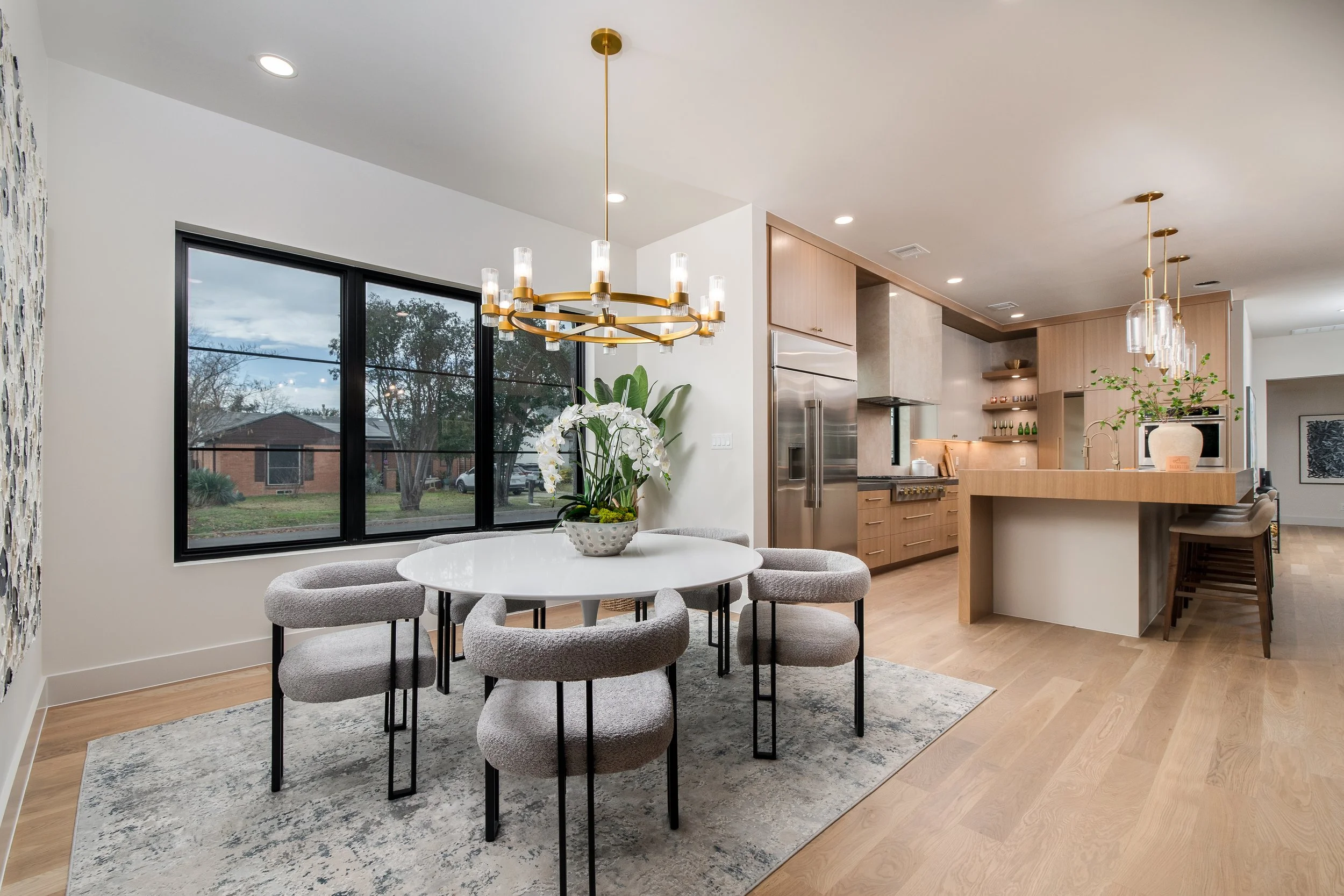 Dining area with a white round table, six upholstered chairs, a potted orchid centerpiece, large window, and modern kitchen in background featuring light wood cabinets, stainless steel refrigerator, and pendant lights.