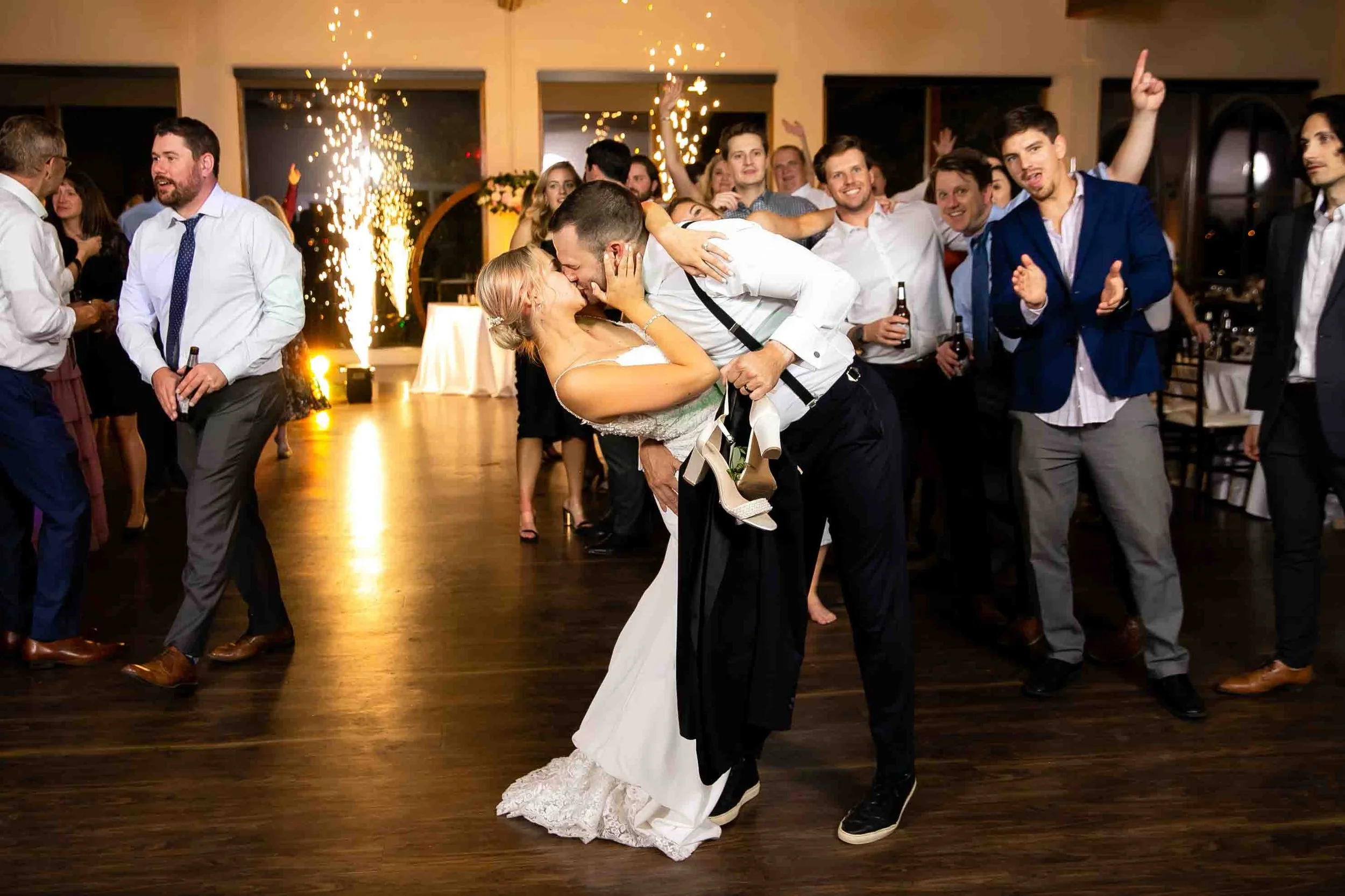 Couple sharing a kiss on the dance floor at a wedding reception, surrounded by smiling guests, with sparklers in the background.