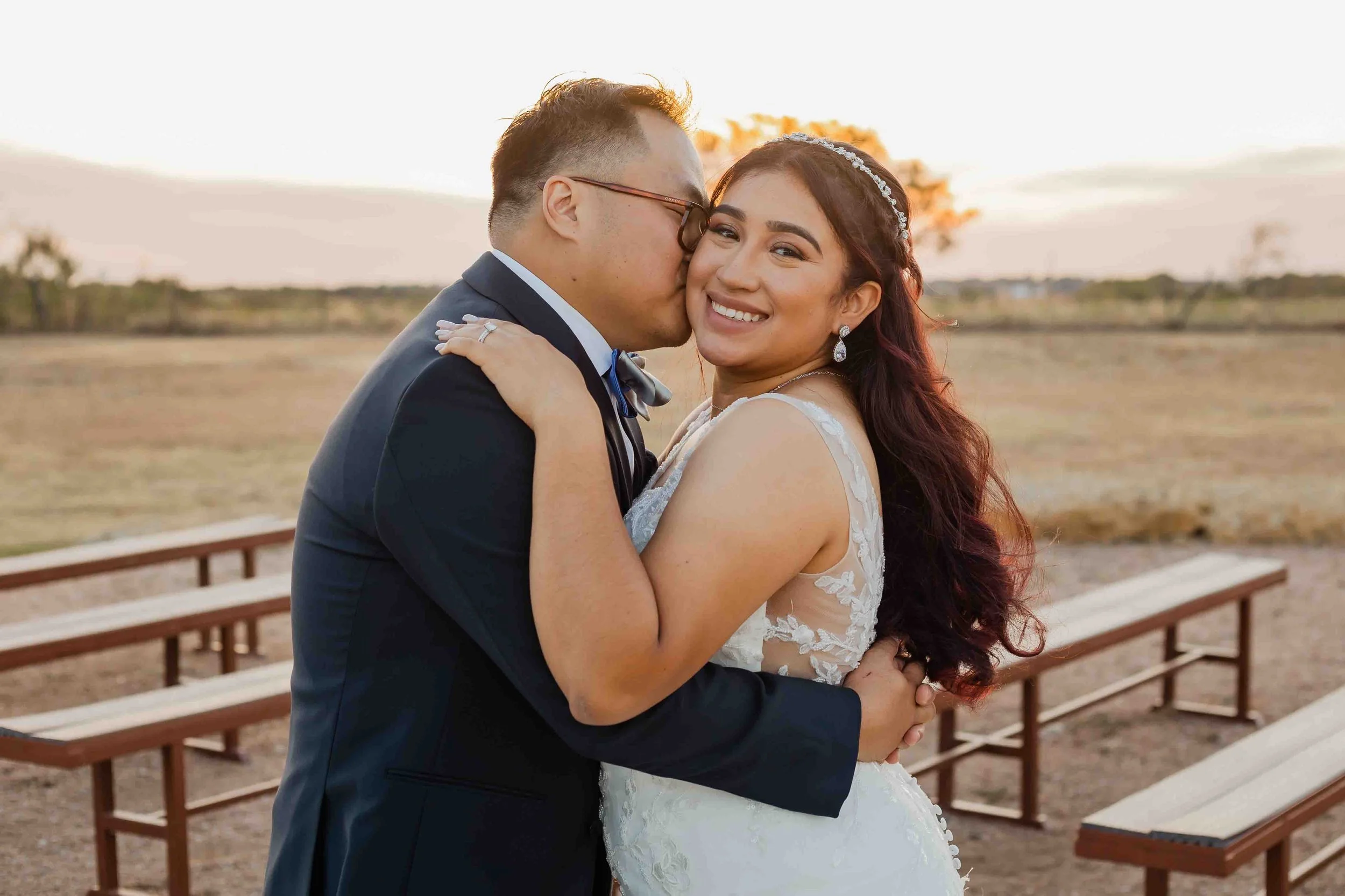 A newlywed couple embraces outdoors during sunset; the groom in a dark suit and glasses kisses the bride, who wears a white lace wedding dress and a tiara, smiling at the camera.