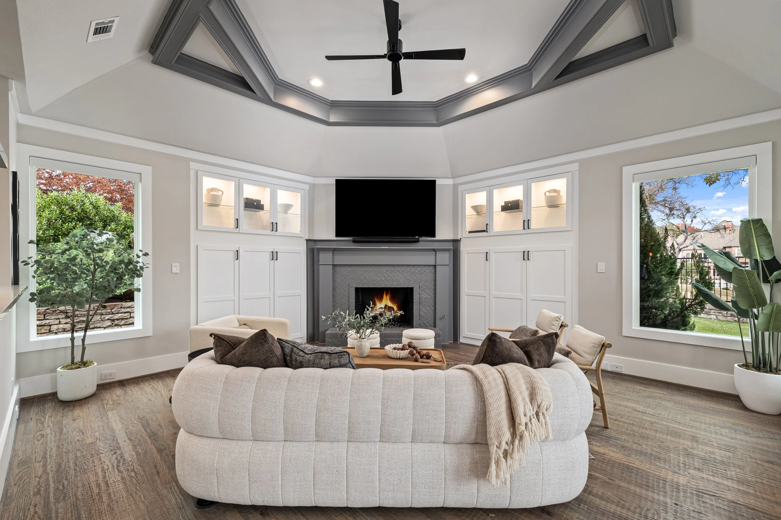 Living room with a round beige sofa, dark brown and beige cushions, a wooden coffee table, a fireplace with a TV above, large windows showing greenery outside, a ceiling fan, built-in white cabinets around the fireplace, and hardwood floors.