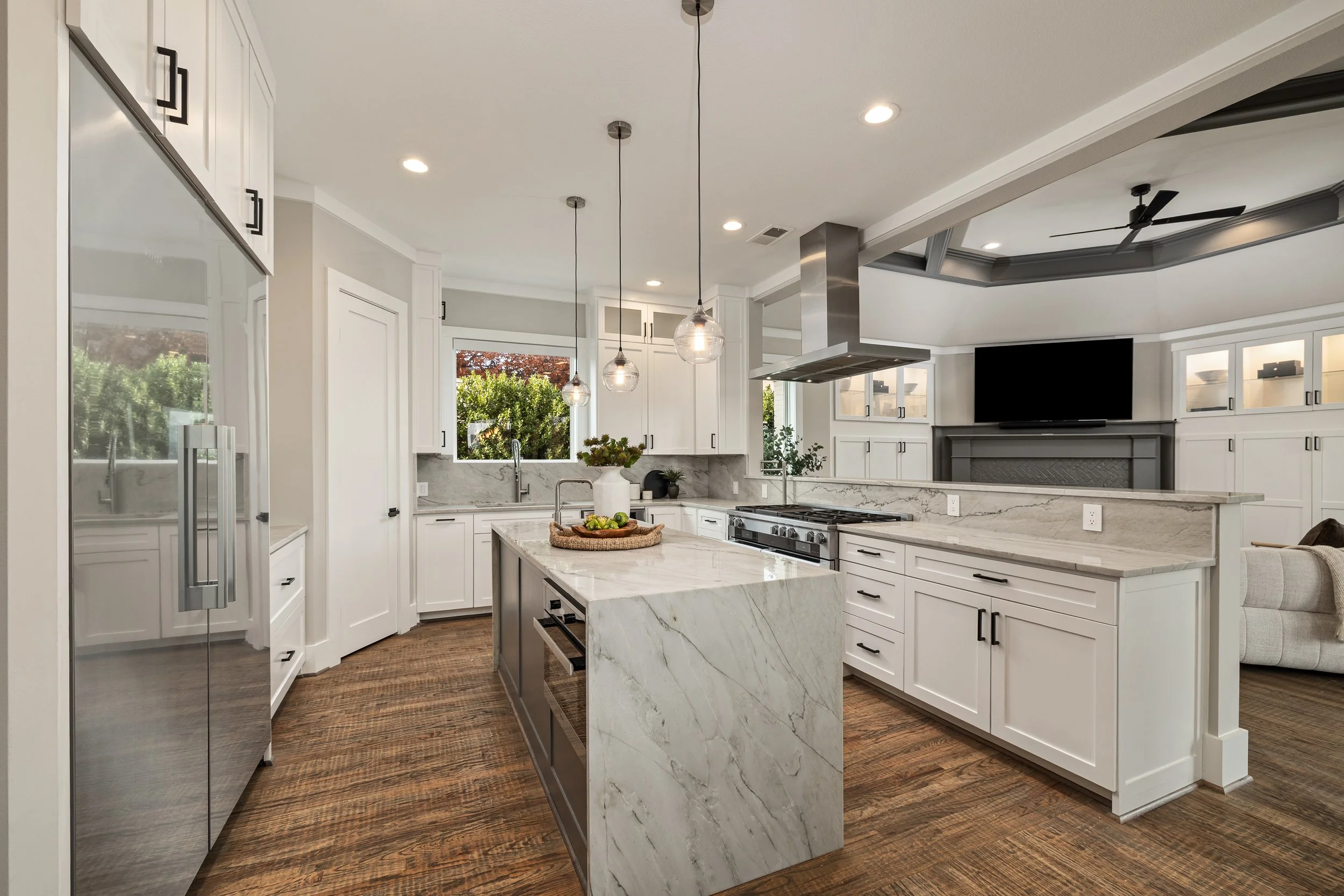 Modern kitchen with white cabinets, a marble island, stainless steel appliances, pendant lights, hardwood floors, and a living room with a TV and fireplace in the background.