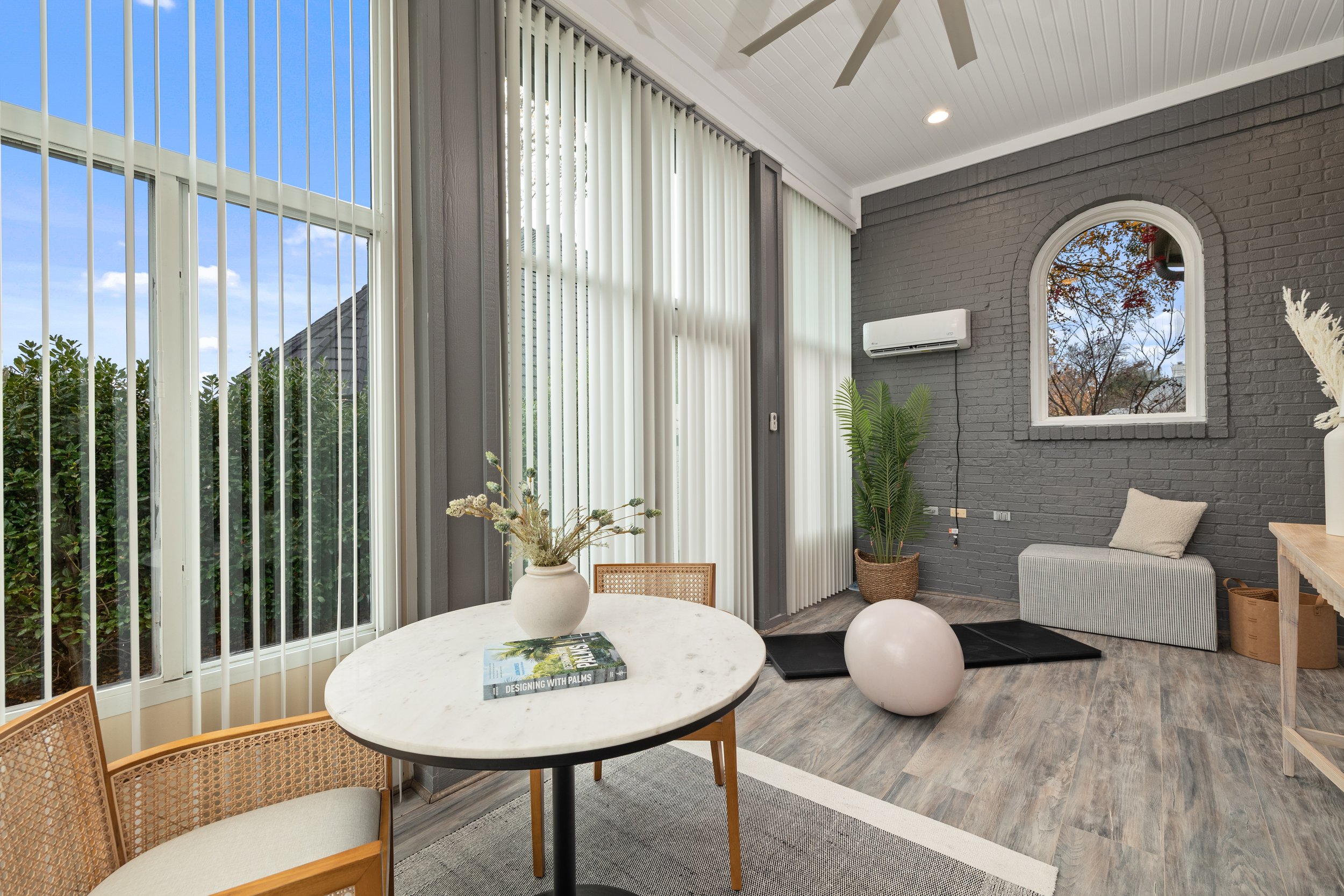 Interior of a modern living room with large windows, gray brick wall, potted plants, a small bench, and decorative items.