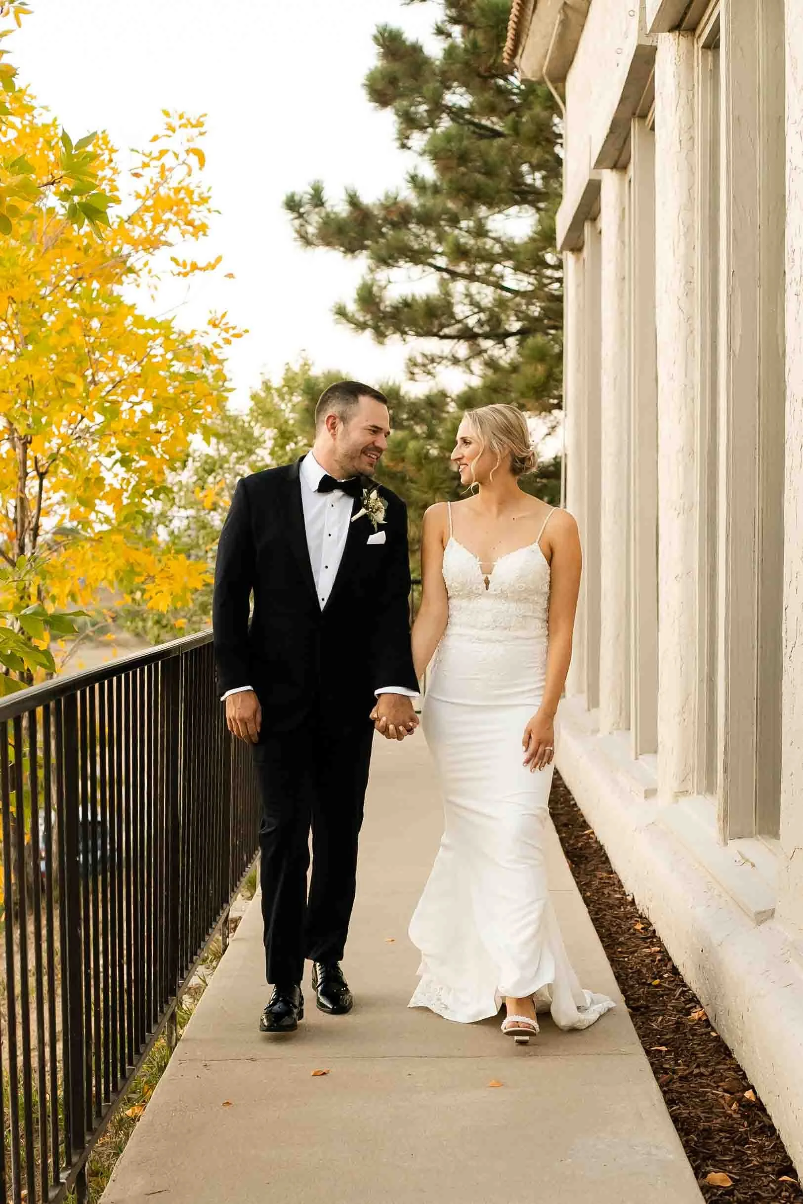 A newlywed couple holding hands walking on a sidewalk with autumn leaves and trees in the background.