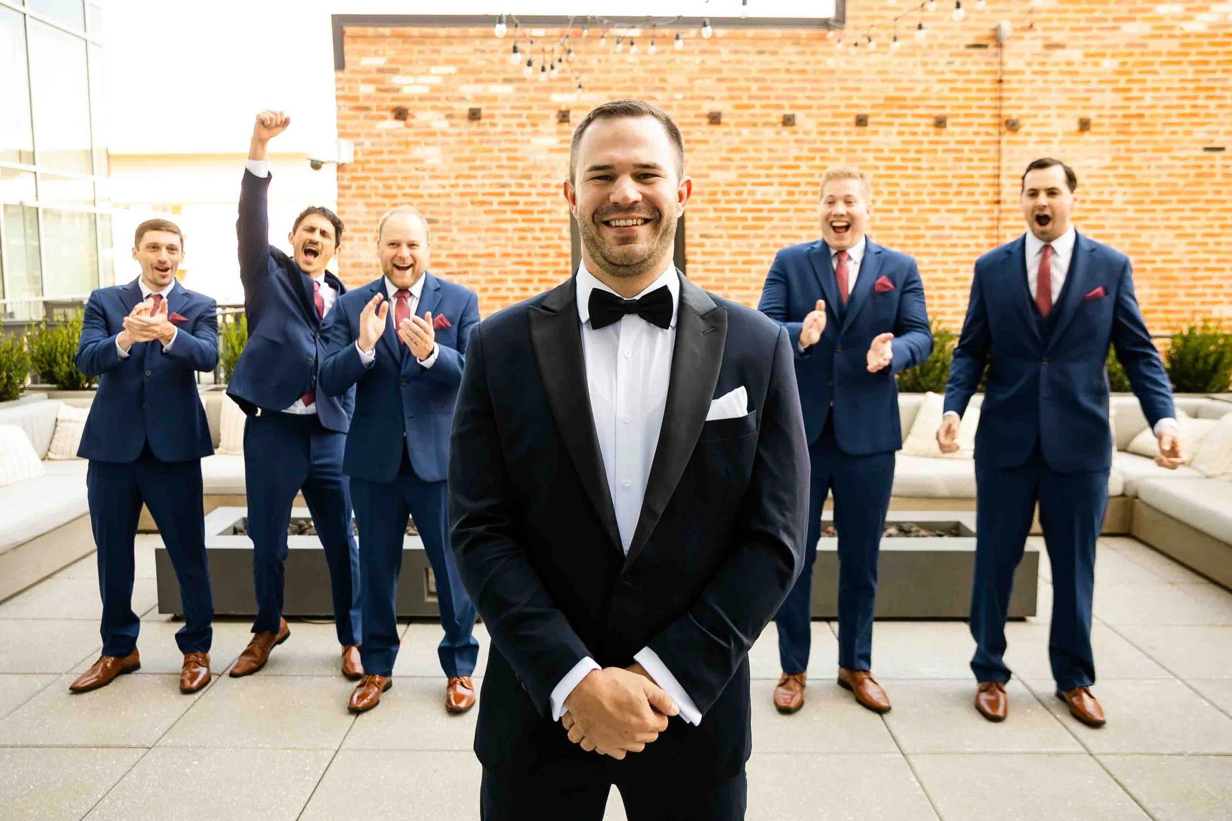 A groom in a tuxedo smiling in front of six groomsmen dressed in matching navy suits and red ties, celebrating on a rooftop patio with a brick wall background and string lights.