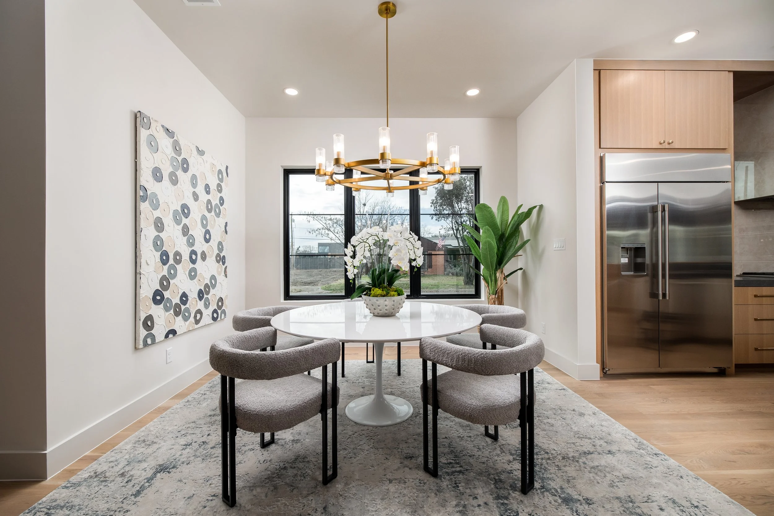 Modern dining room with a white round table, six cushioned chairs, a large window, a decorative chandelier, botanical artwork on the wall, and a kitchen with stainless steel appliances and wooden cabinetry in the background.