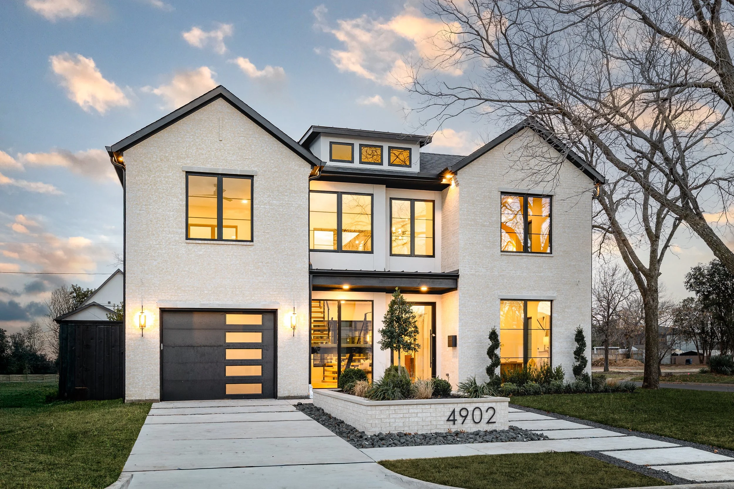 Modern two-story house with white brick exterior, black garage door, and large windows, illuminated from within at dusk, with a landscaped front yard and a tree on the right side.