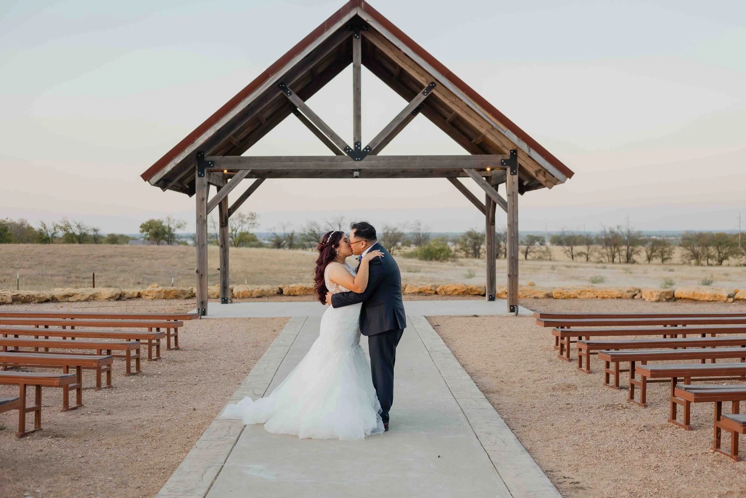 A bride and groom kissing under a wooden pavilion at an outdoor wedding venue in a desert landscape.