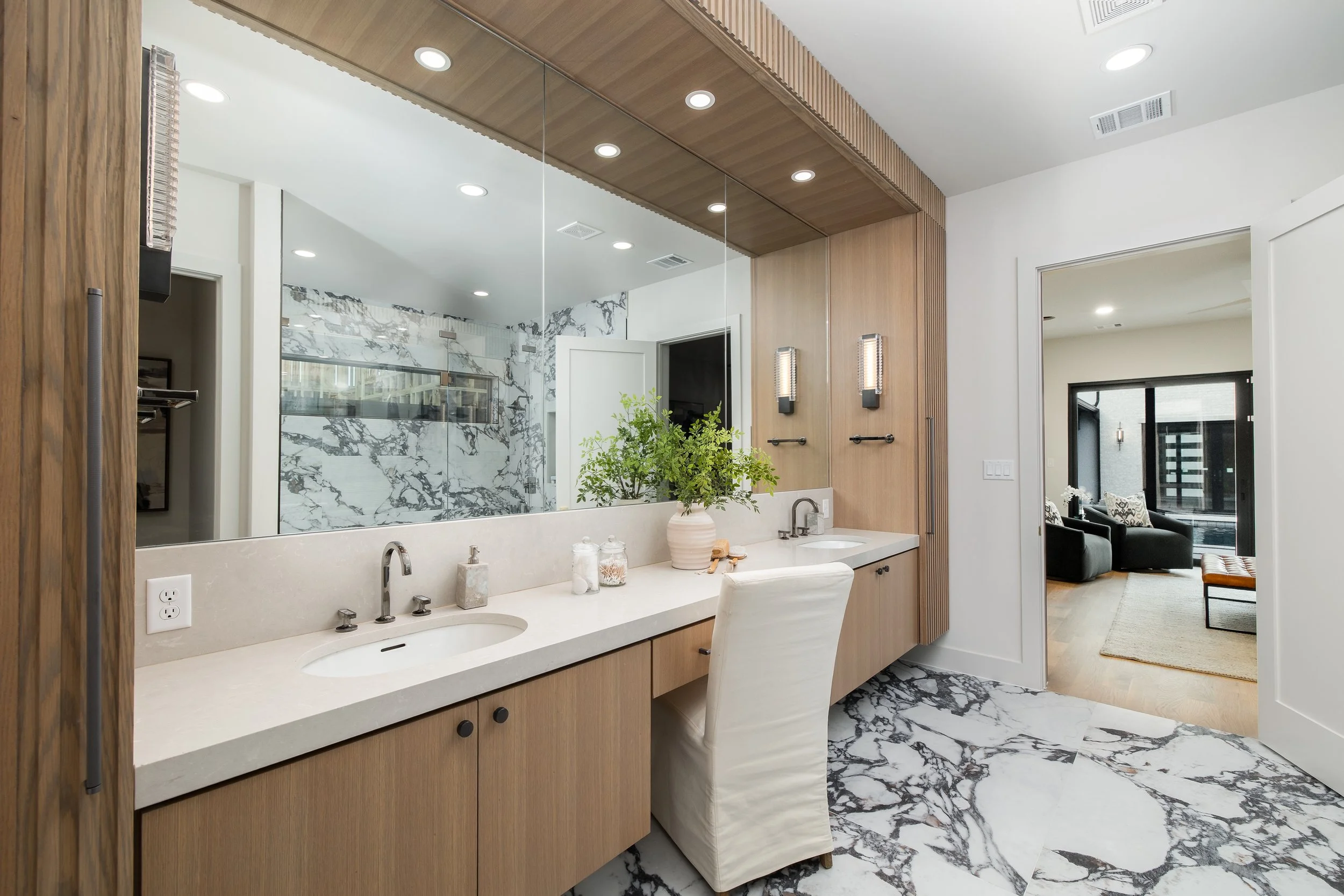 Modern bathroom with dual sinks, marble flooring, wooden cabinetry, and a large mirror. An adjoining room with black furniture and a sliding glass door is visible.