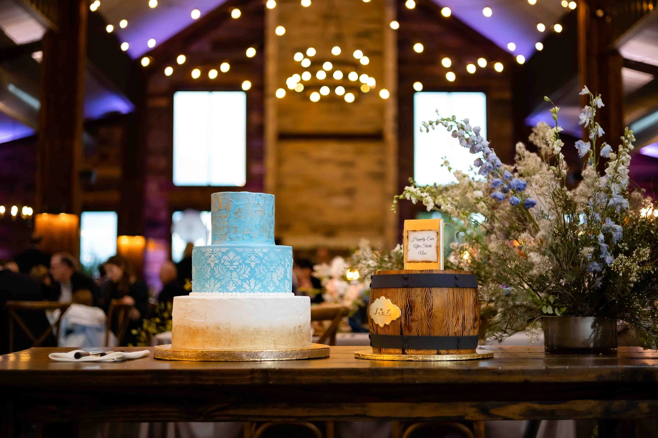 A three-tiered wedding cake with a white bottom tier, a light blue middle tier with a damask pattern, and a blue top tier, set on a wooden table. Next to the cake is a small wooden barrel with a framed note on top. There is a large floral arrangement