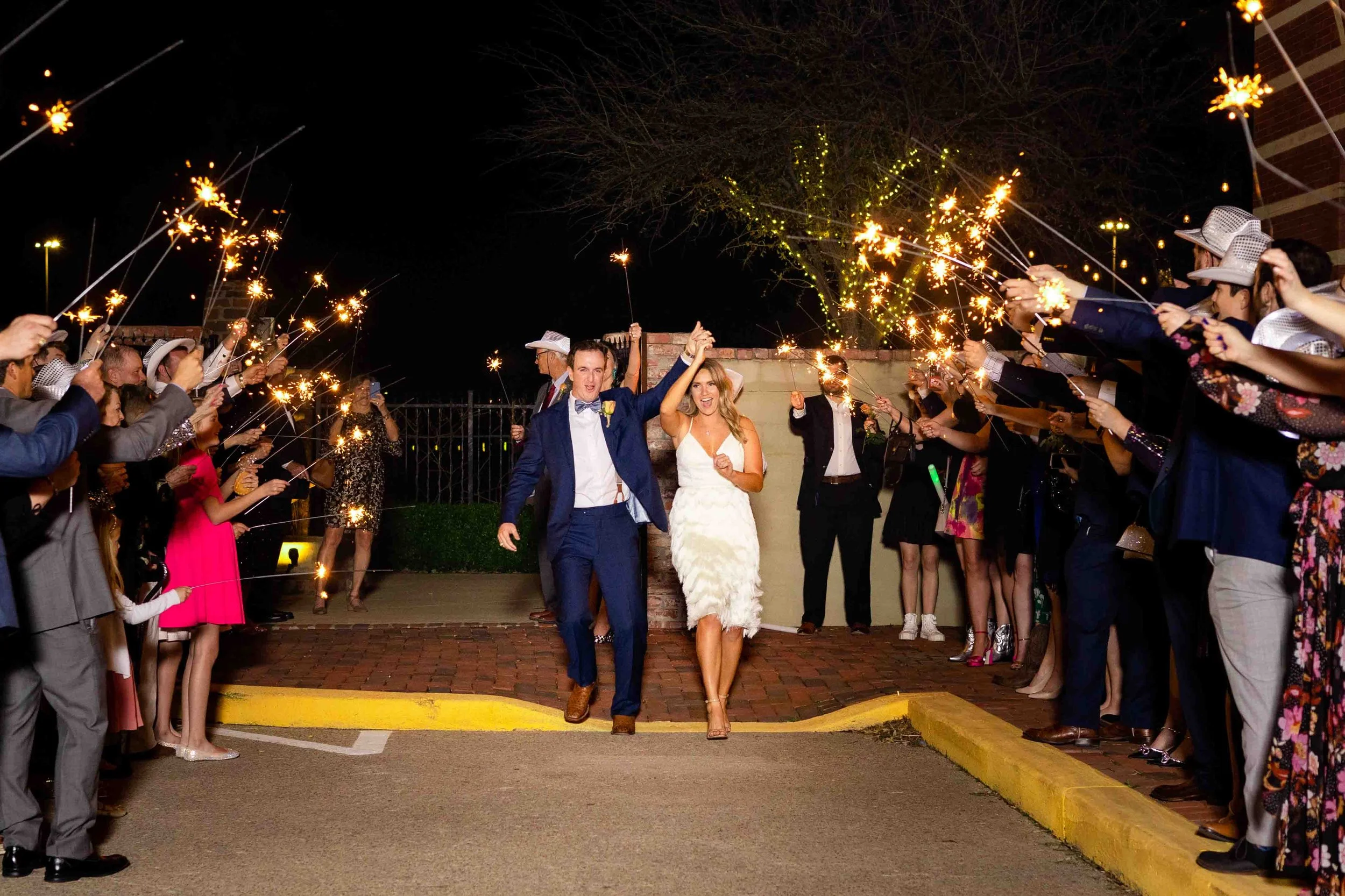 A newlywed couple walking through a sparklers celebration at night, surrounded by friends and family.