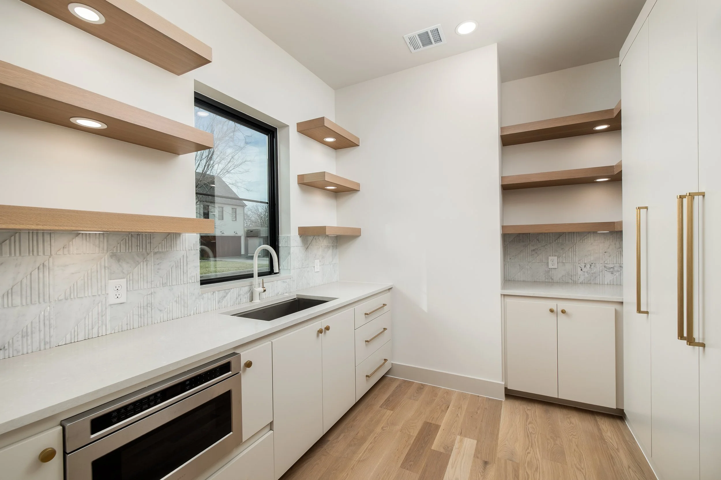 Modern kitchen with white cabinets, wooden open shelves, a window above the sink, and a microwave oven built into the lower cabinets.